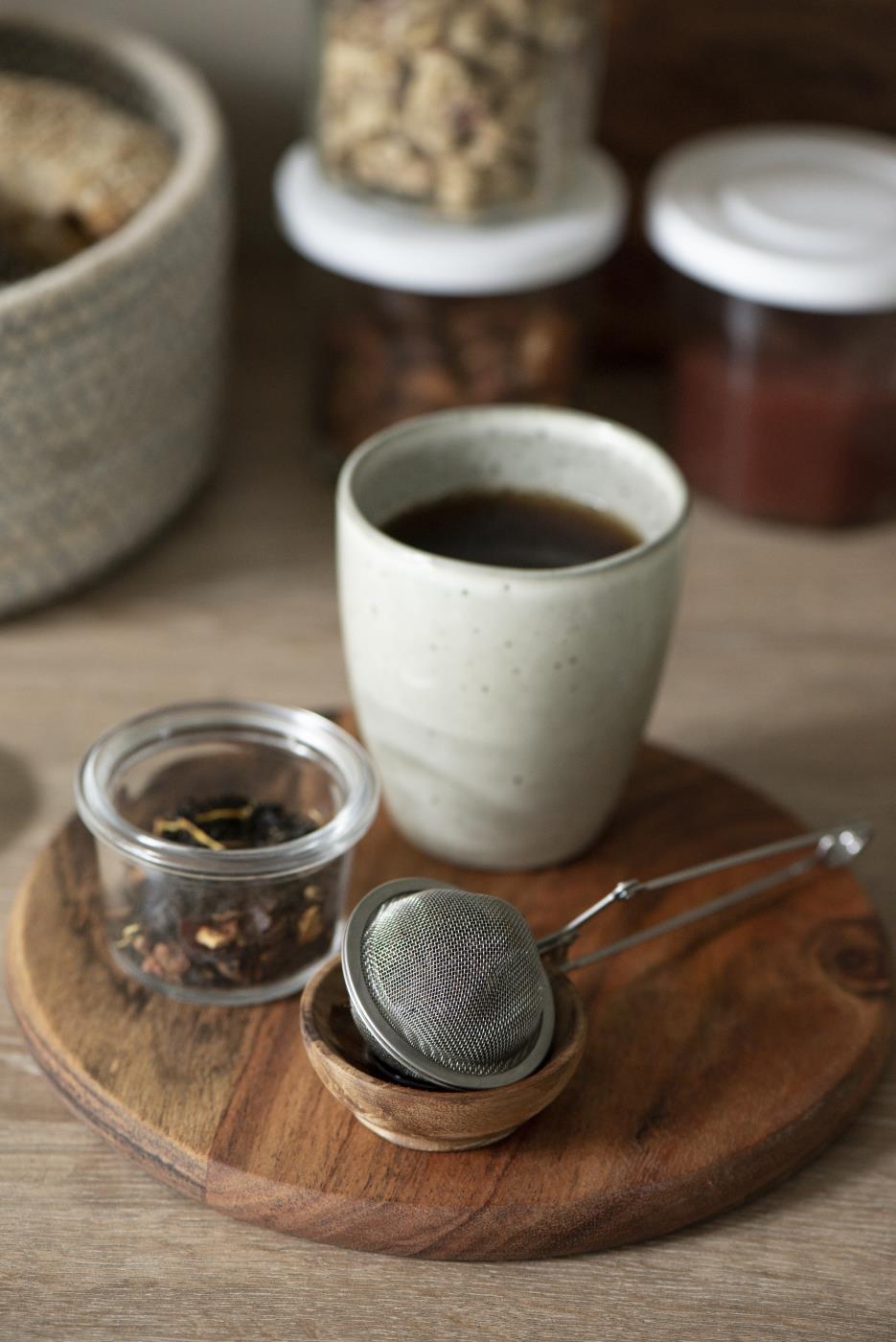 Eine Tasse Kaffee steht auf einem Holzbrett, daneben ein Glas mit Teeblättern und ein Teesieb in einer kleinen Schale.