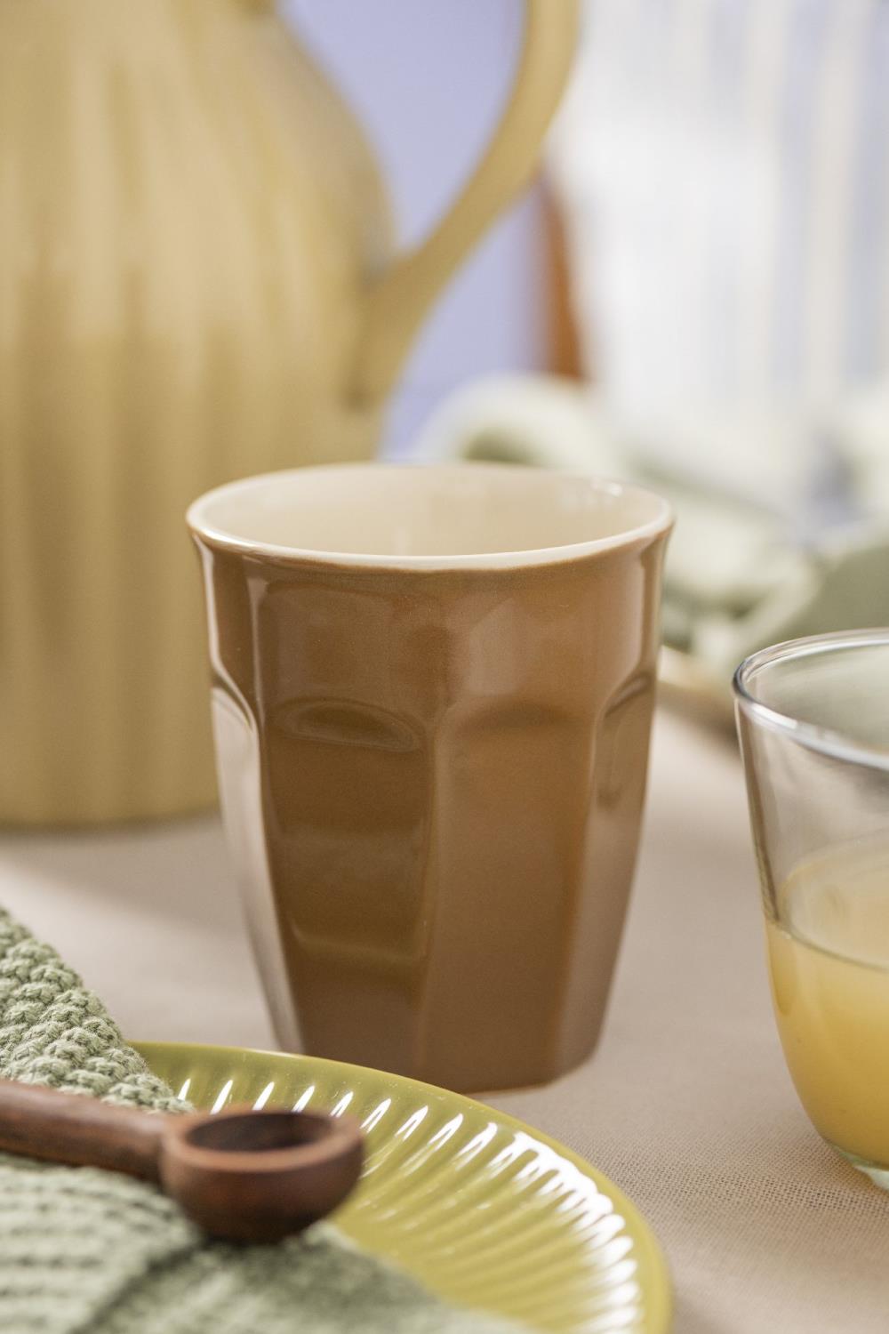 Brauner Becher steht auf einem Tisch, umgeben von einem Krug, einem Glas, einem Teller und einem Lappen in sanften Farben.