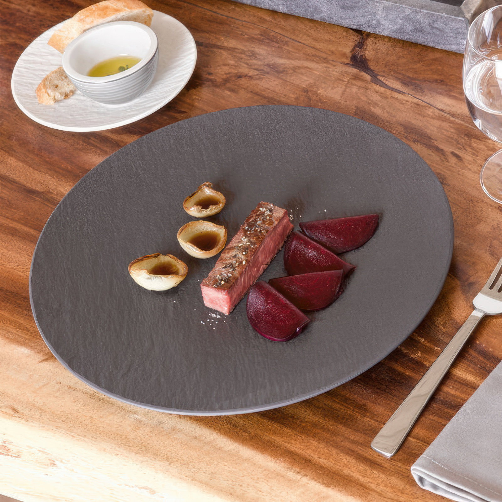 Ein grauer Teller mit einem Stück Fleisch, Rote-Bete-Scheiben und kleinen Zwiebeln. Im Hintergrund Brot und Öl in einer Schale.