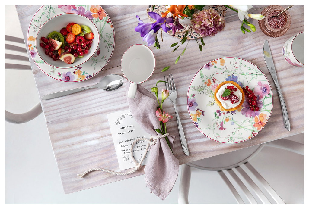Tischgedeck mit buntem Obstsalat, einem Dessert, Besteck, einer Serviette und einer Blumenvase. Hand
