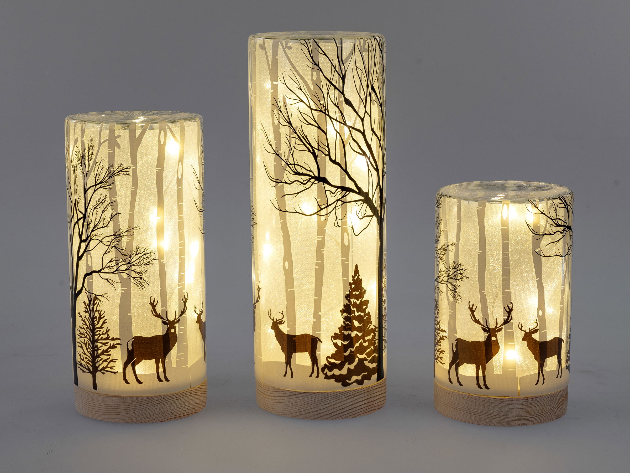 Drei beleuchtete Kerzen mit winterlichen Motiven: Bäume, Rehe und einen Tannenbaum auf transparentem