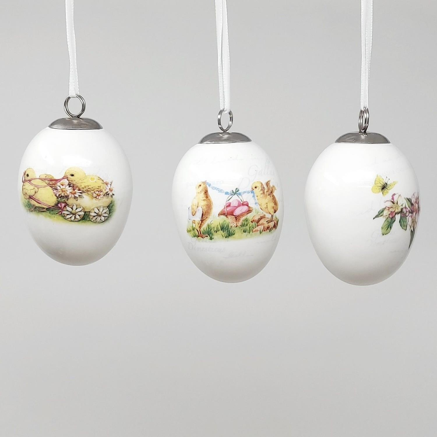 Drei hängende Ostereier aus Porzellan mit bunten, frühlingshaften Motiven von Küken und Blumen.
