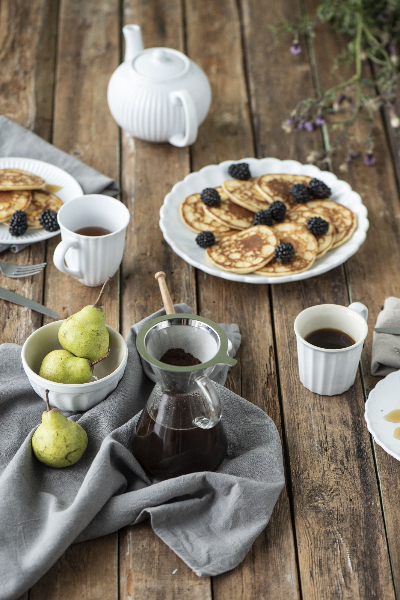Tischgedeck mit Pfannkuchen, Brombeeren, Birnen, Kaffeekanne und Tassen auf einem Holztisch, umgeben von einem grauen Tuch.
