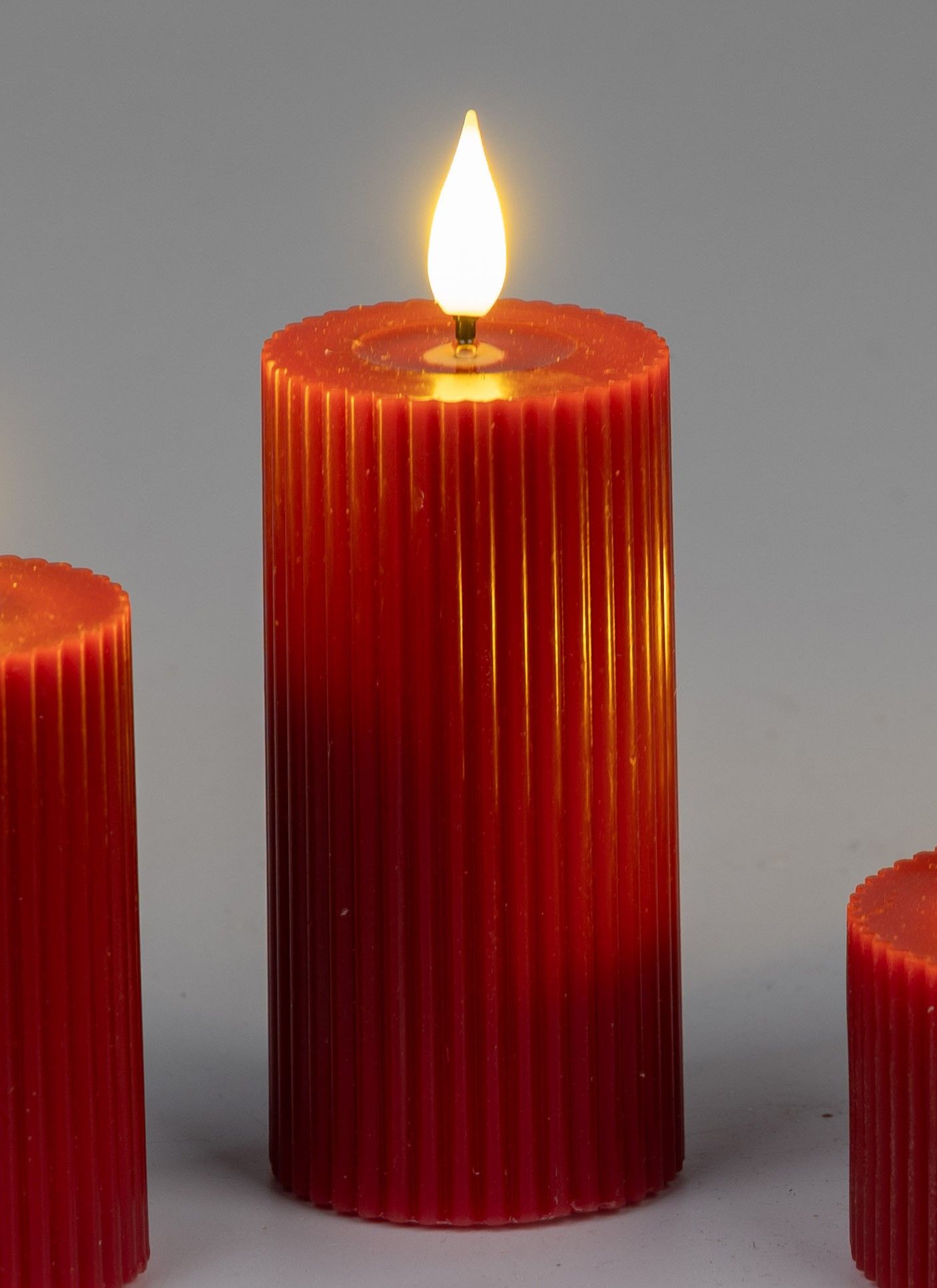 Drei rote Kerzen mit gerillter Oberfläche, eine brennt mit einer gelben Flamme, vor neutralem Hinter