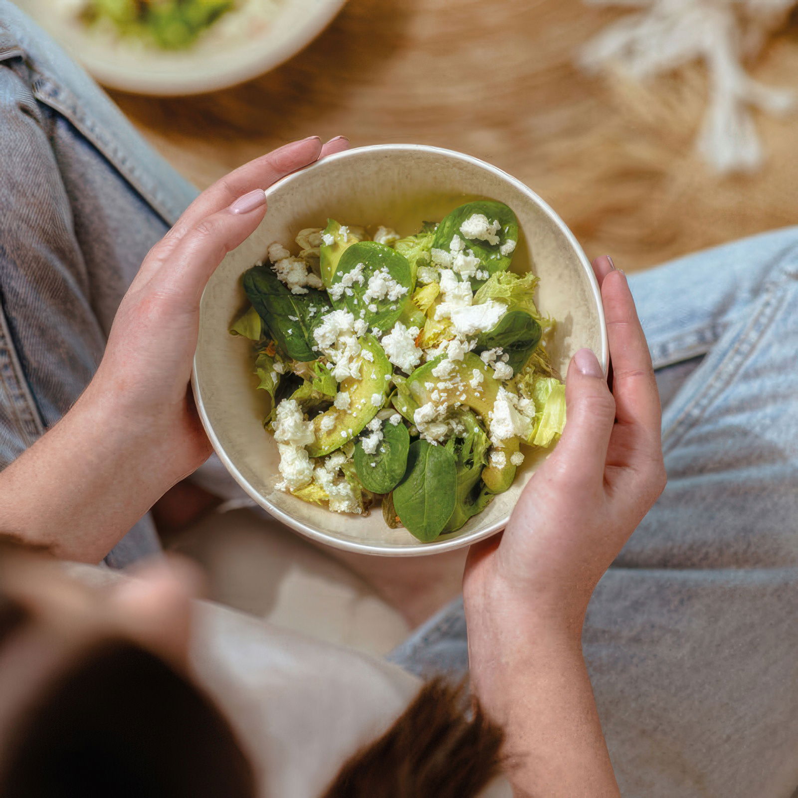 Eine Person hält eine Schüssel mit grünem Salat, garniert mit Feta-Käse, während sie auf einem Teppi
