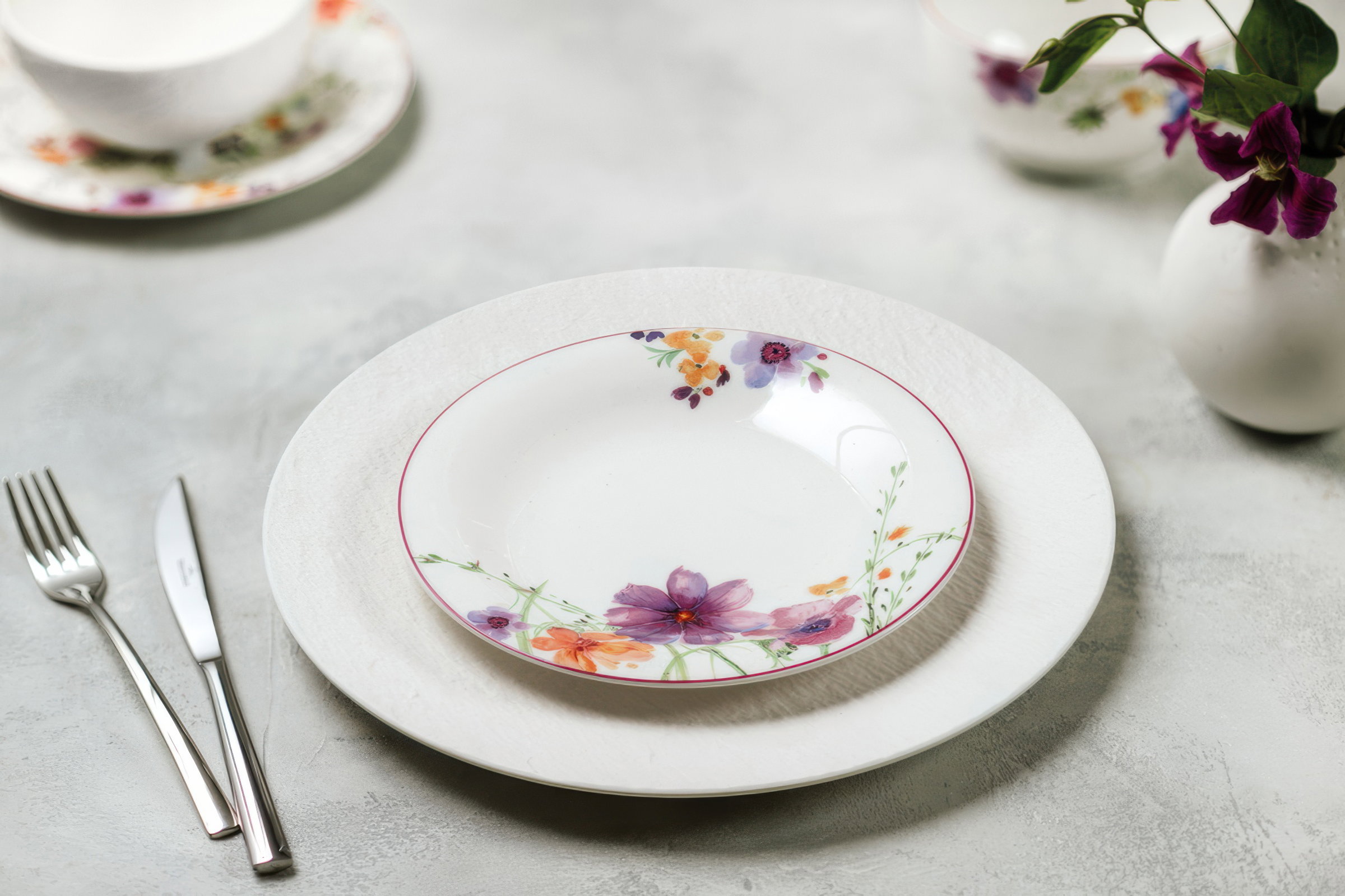 Ein gedeckter Tisch mit einem weißen Teller, einem flachen Teller mit Blumenmuster, Besteck und einer kleinen Blumenvase.