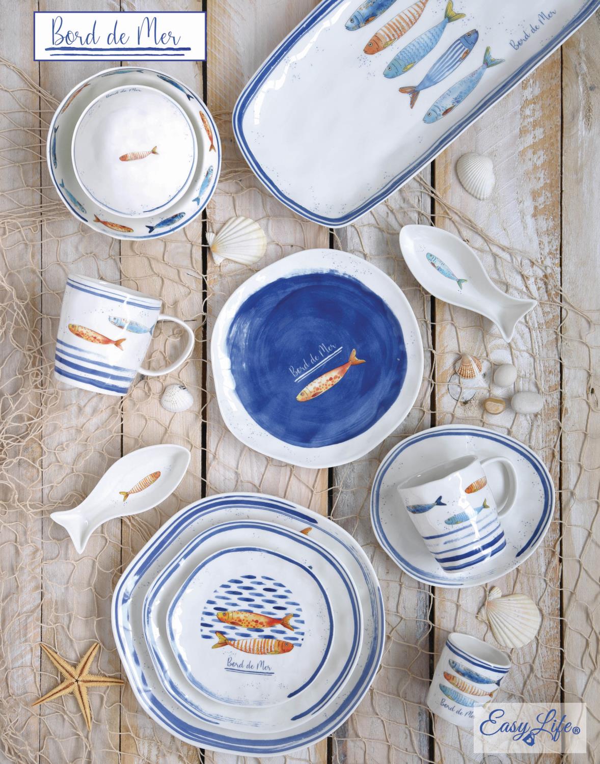 Teller, Tassen und Schalen mit Fischmotiven in Blau und Weiß auf einem Holzuntergrund, umgeben von Muscheln und Netz.