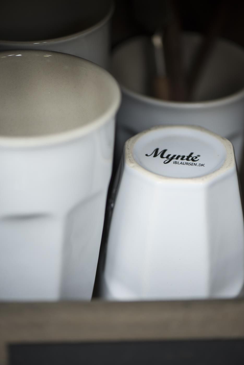 Weißes Geschirr in einem Schrank, darunter eine Tasse mit dem Schriftzug "Mynte" und weiteren Tassen im Hintergrund.