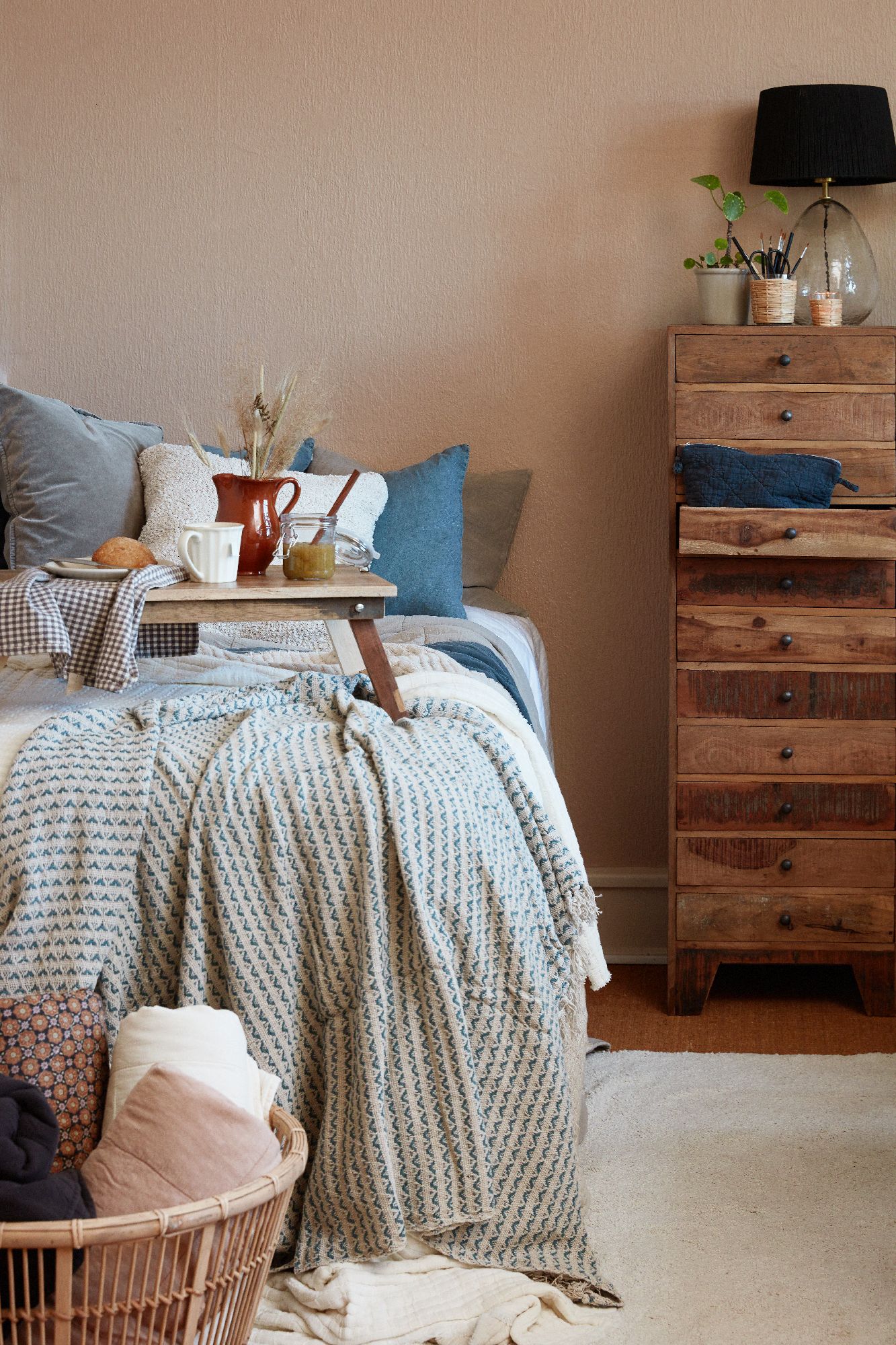 Ein Schlafzimmer mit einem Bett, einer Holzkommode, einem Nachttisch und verschiedenen Textilien in neutralen Farben.