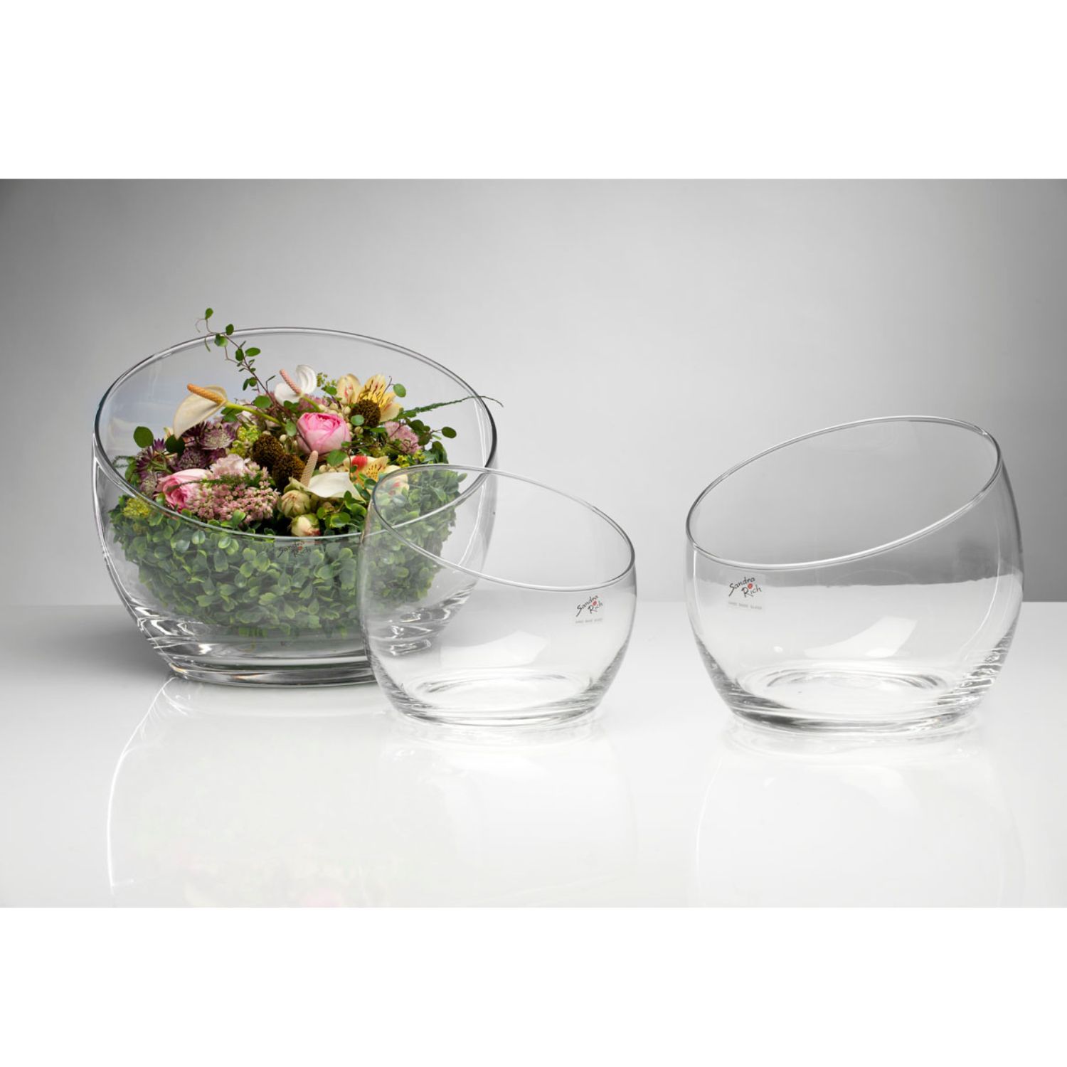 Drei transparente Glasschalen in verschiedenen Größen, eine mit Blumen und Grünpflanzen gefüllt, auf einem hellen Untergrund.