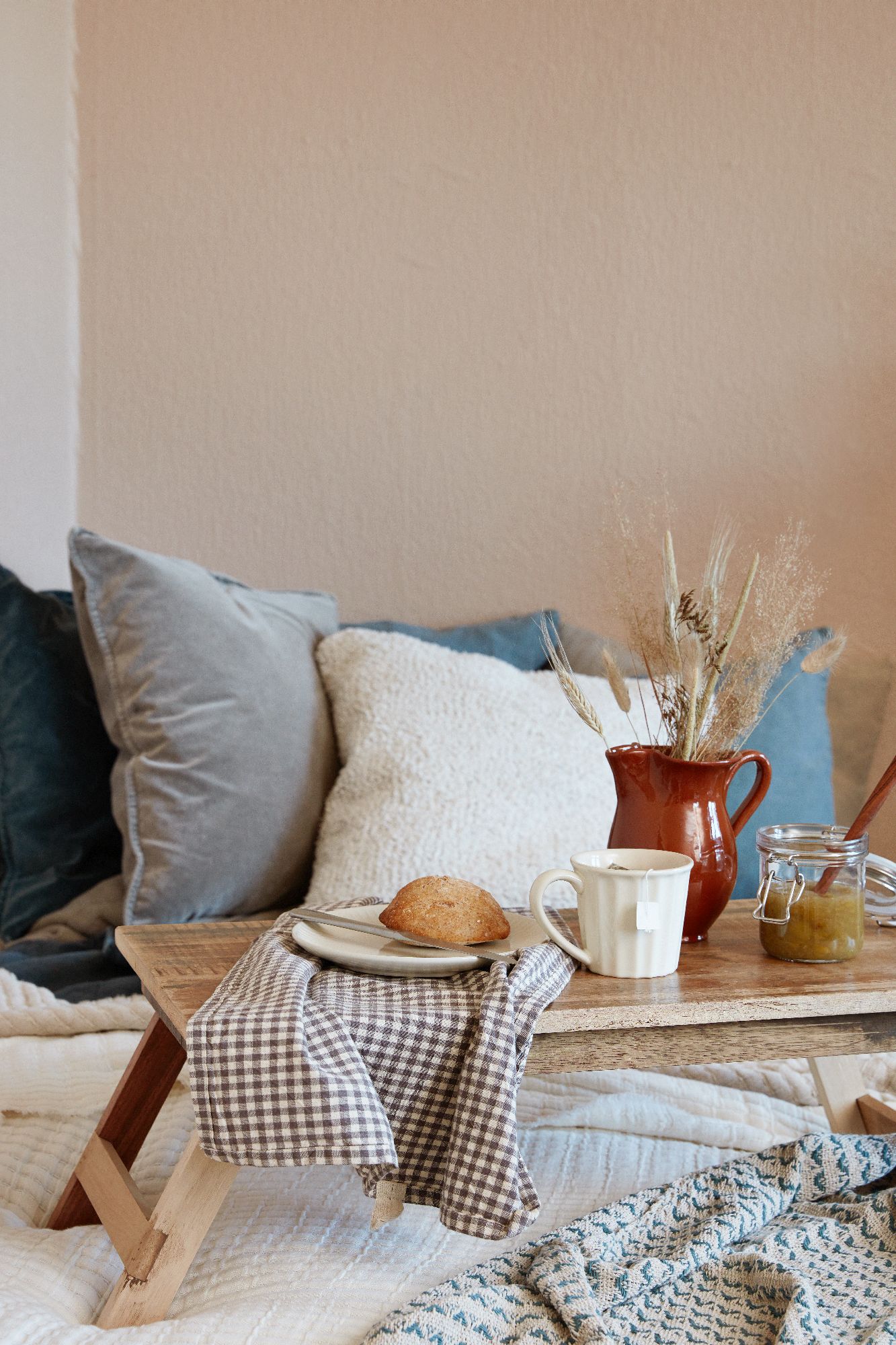 Ein Holztisch mit einem Teller, einer Tasse, einem Brötchen, einem Krug und einem Glas auf einer Decke, umgeben von Kissen.