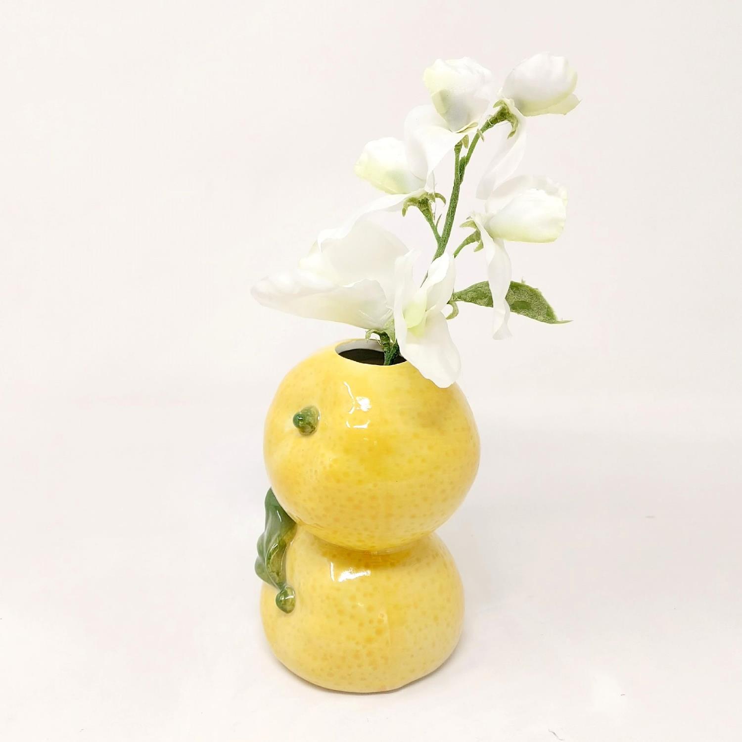 Gelbe, runde Vase in Form von zwei übereinanderliegenden Zitrusfrüchten mit weißen Blumen und grünen Blättern.