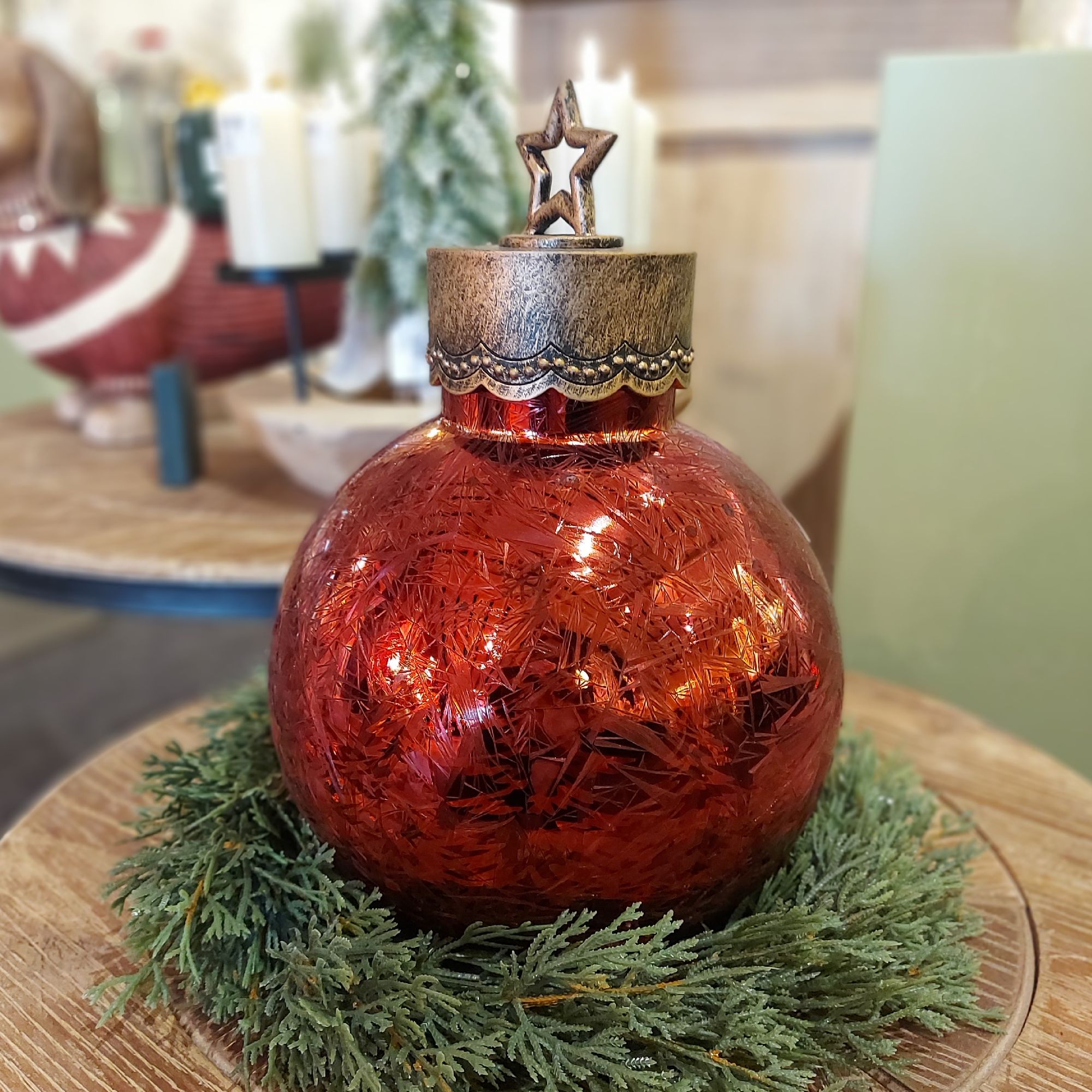 Rote Weihnachtskugel mit glitzernder Oberfläche und Sternenaufsatz, umgeben von einem grünen Tannenz