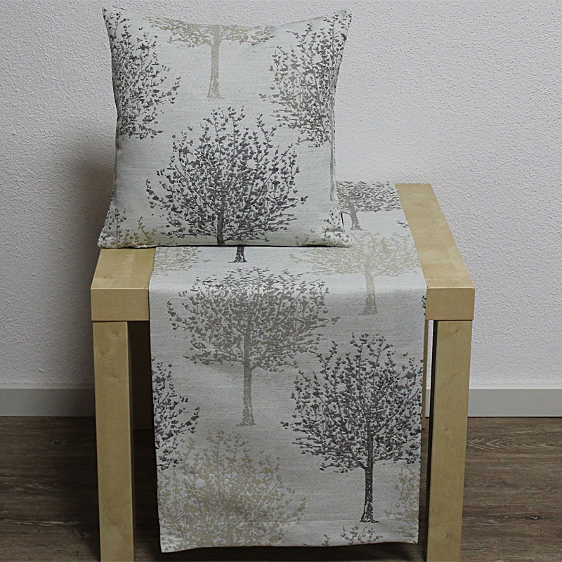 Ein Kissen und ein Tischläufer mit Baum-Muster auf einem hellen Tisch vor einer neutralen Wand.
