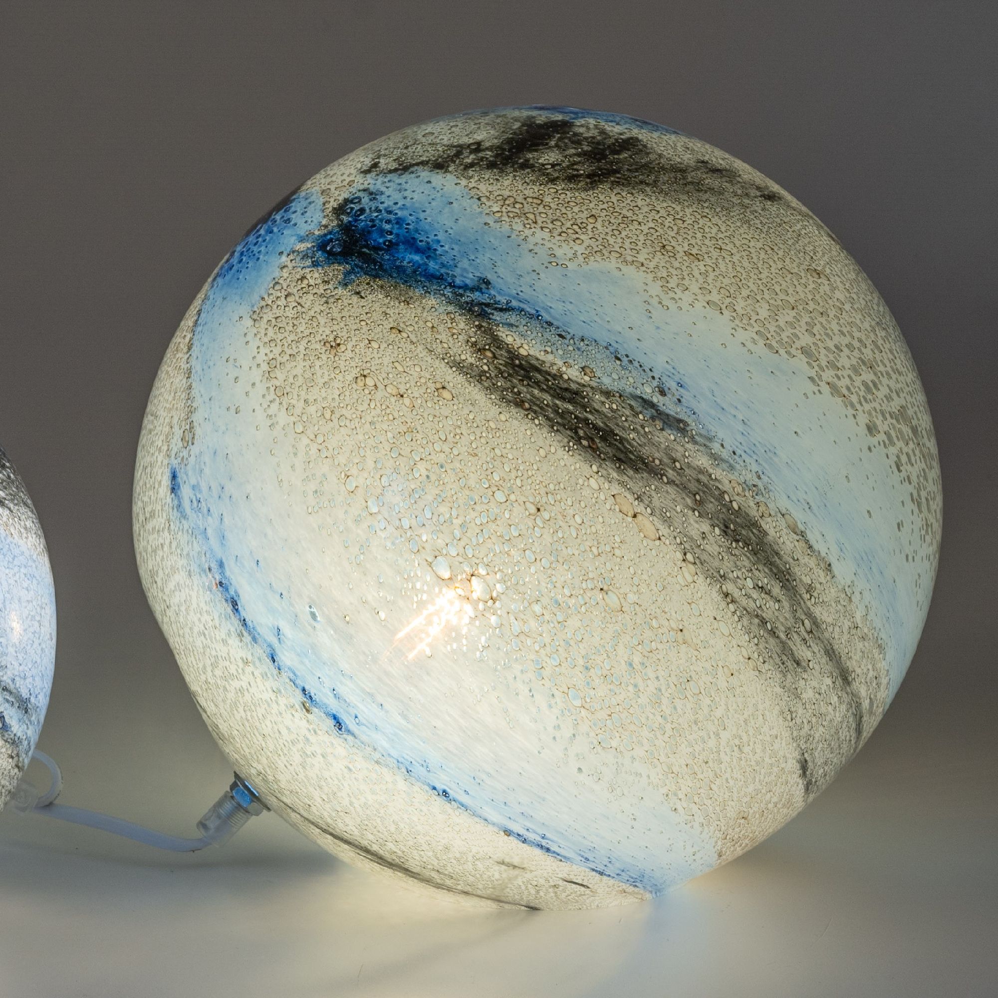 Glaslampe in Kugelform mit blauen und grauen Farbverläufen sowie Blasenstruktur auf der Oberfläche. Beleuchtet von innen.