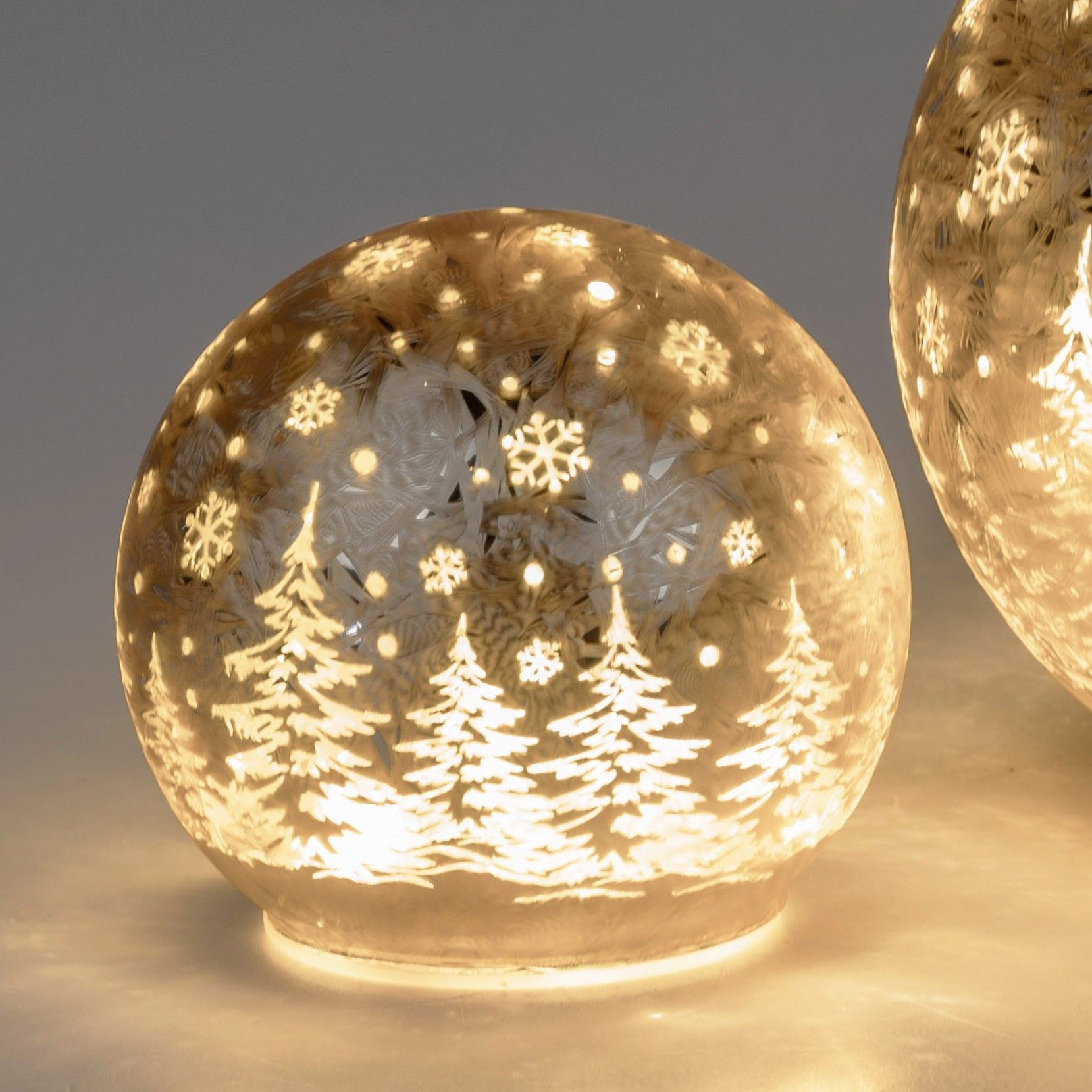 Leuchtende Glaskugel mit winterlichen Motiven, darunter Tannenbäume und Schneeflocken, auf hellem Hi