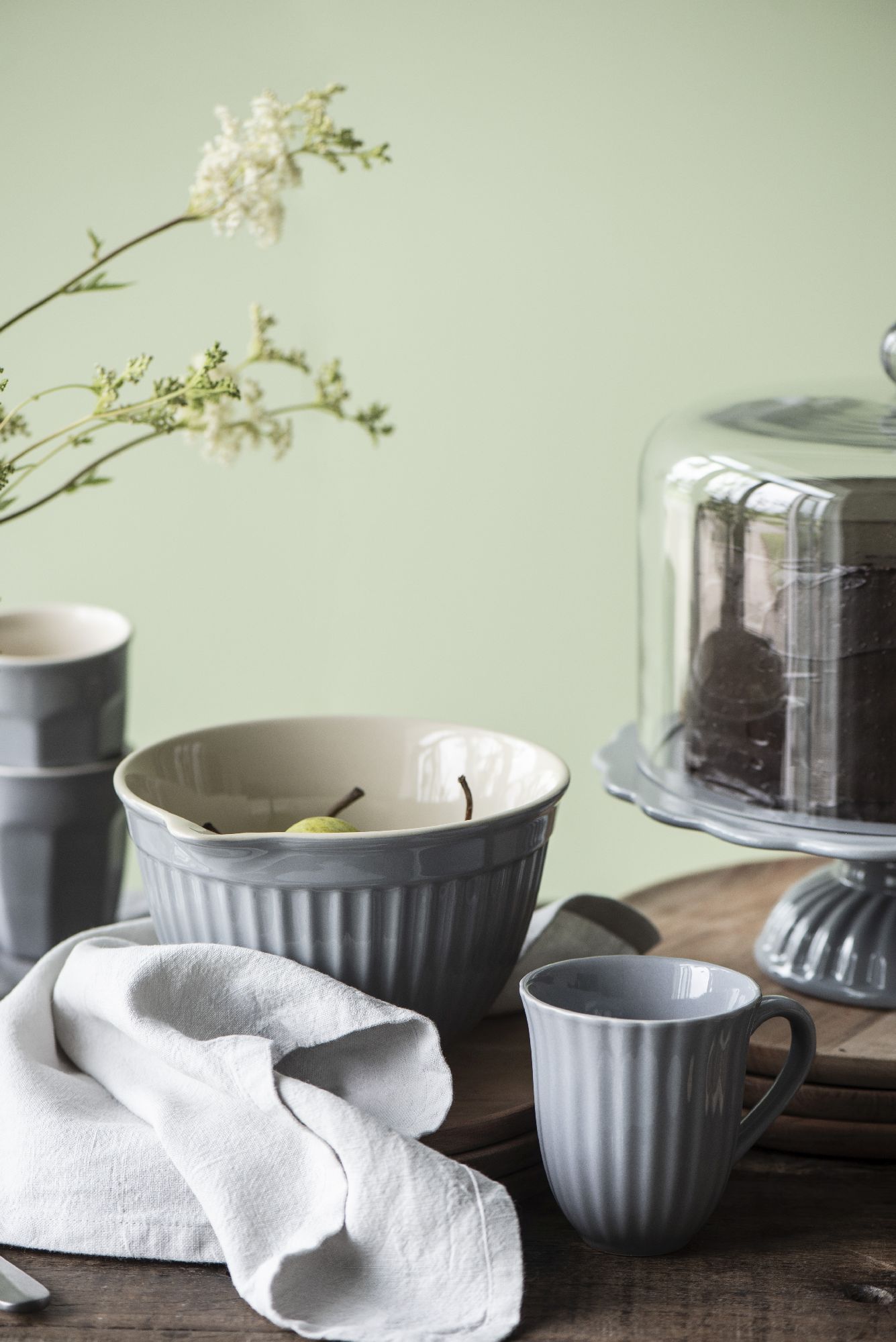 Tischgedeck mit grauen Schalen, Tassen, einem weißen Tuch und einer Glasglocke über einem Kuchen vor grünem Hintergrund.