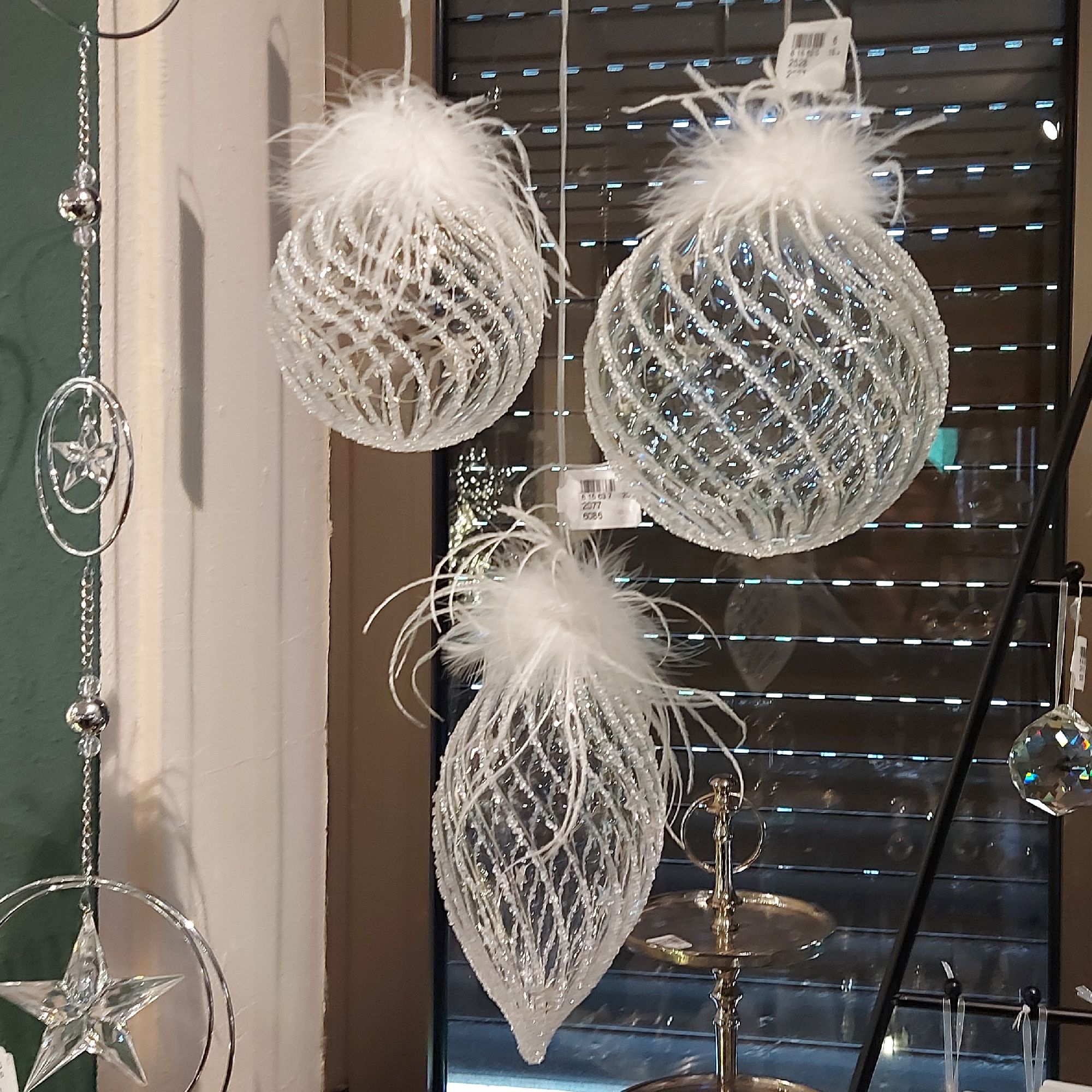 Drei transparente, glitzernde Weihnachtskugeln mit weißen Federn hängen vor einem Fenster. Im Hinter