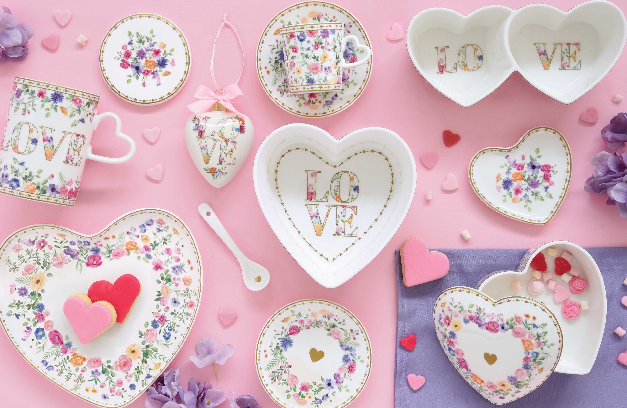 Bunte Geschirrteile in Herzform, Tassen und Dekorationen auf pinkem Hintergrund, mit Blumenmustern und dem Wort "LOVE".