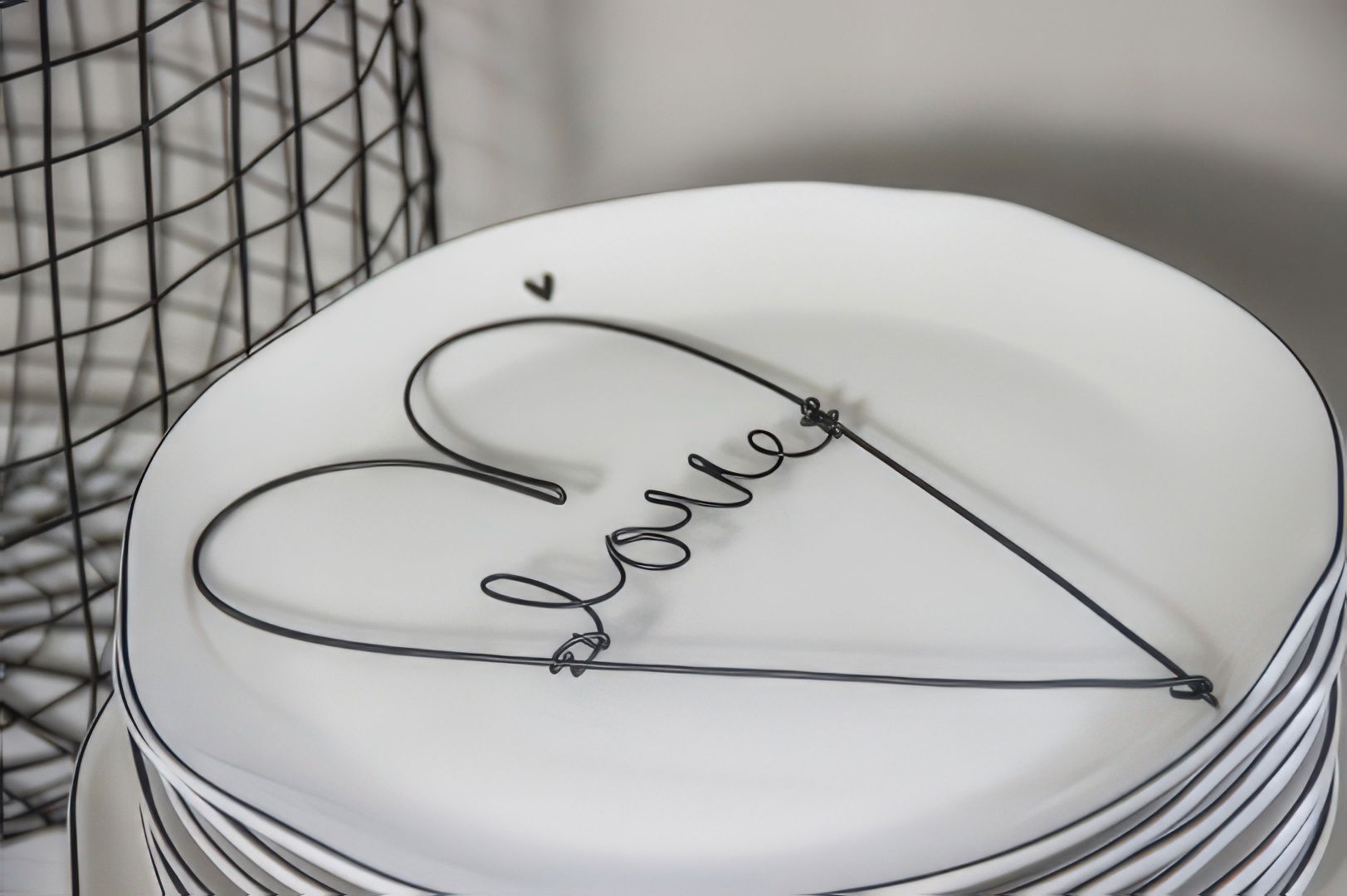 Ein Stapel weißer Teller mit einer schwarzen Drahtschrift, die das Wort „love“ in Herzform bildet. Im Hintergrund ein Drahtkorb.