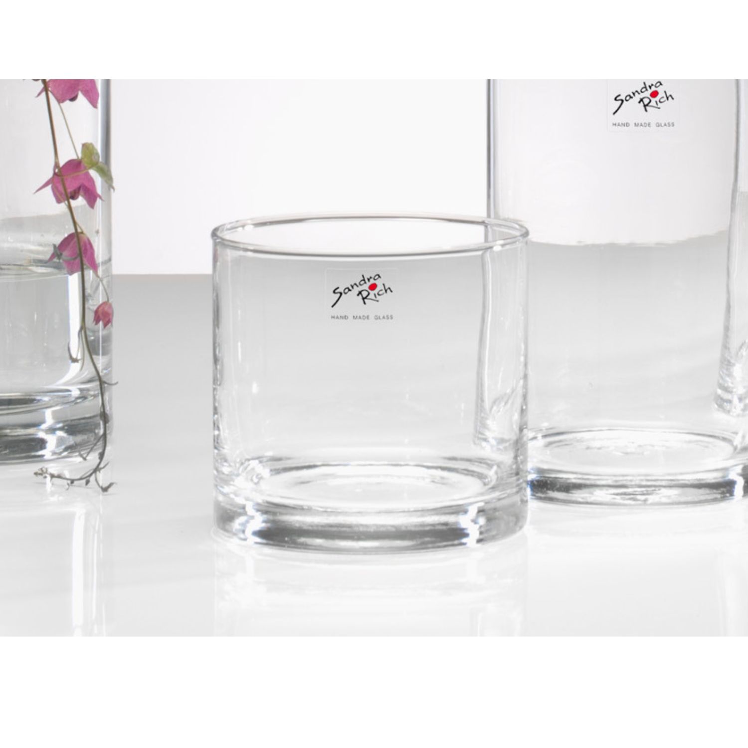 Leeres zylindrisches Glas auf weißem Untergrund, daneben weitere Gläser und ein Blütenstiel