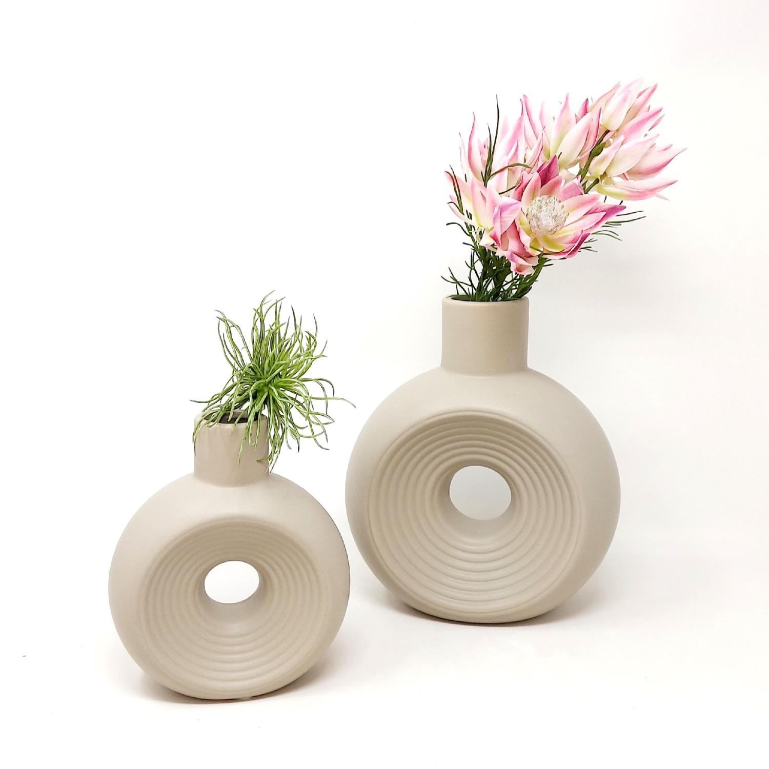 Zwei runde, beige Vasen mit unterschiedlichen Höhen, gefüllt mit Pflanzen und Blumen, vor einem weißen Hintergrund.