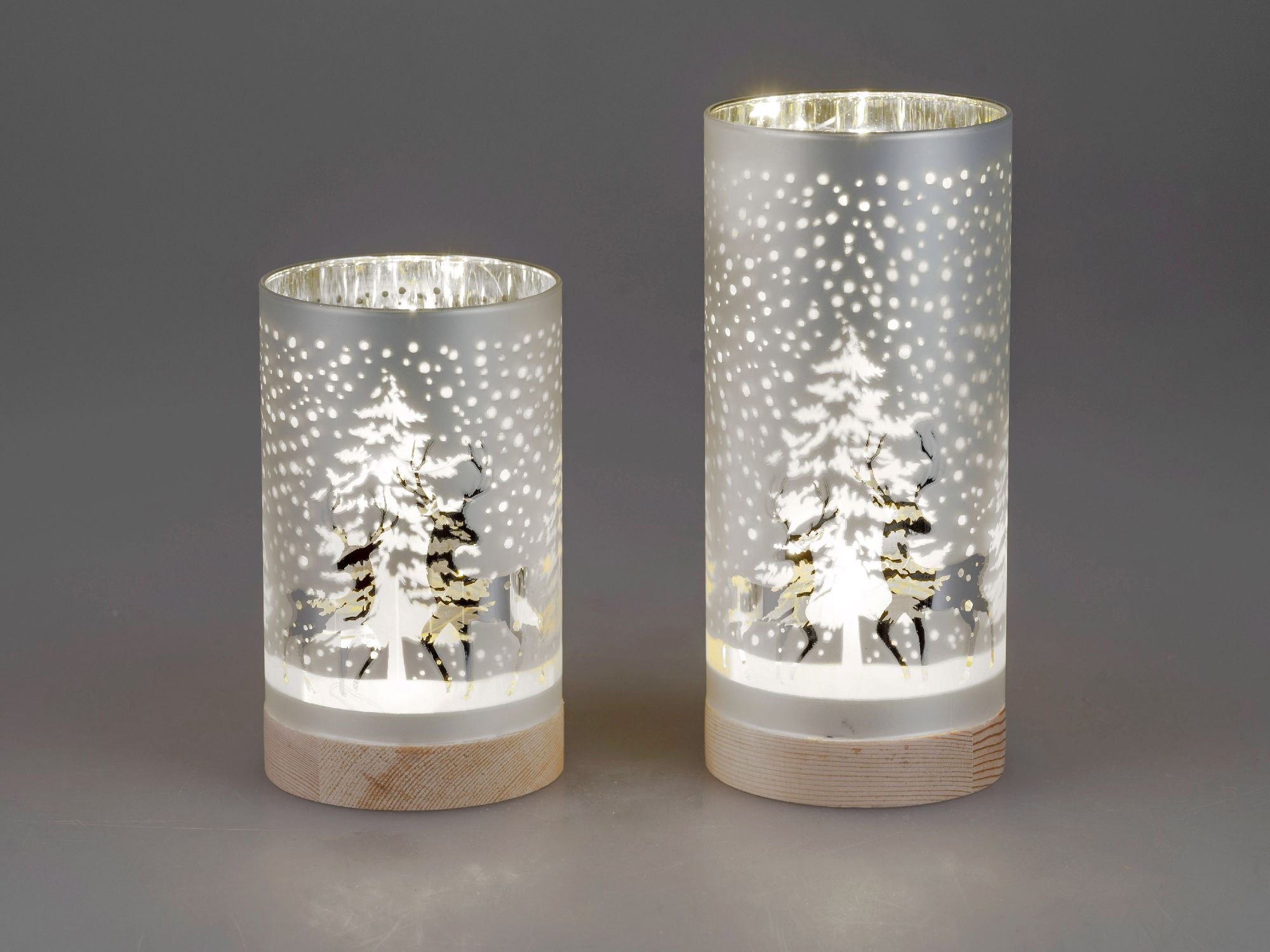 Zwei beleuchtete Windlichter aus Glas mit winterlichen Motiven, umgeben von Schneeflocken, auf Holzs