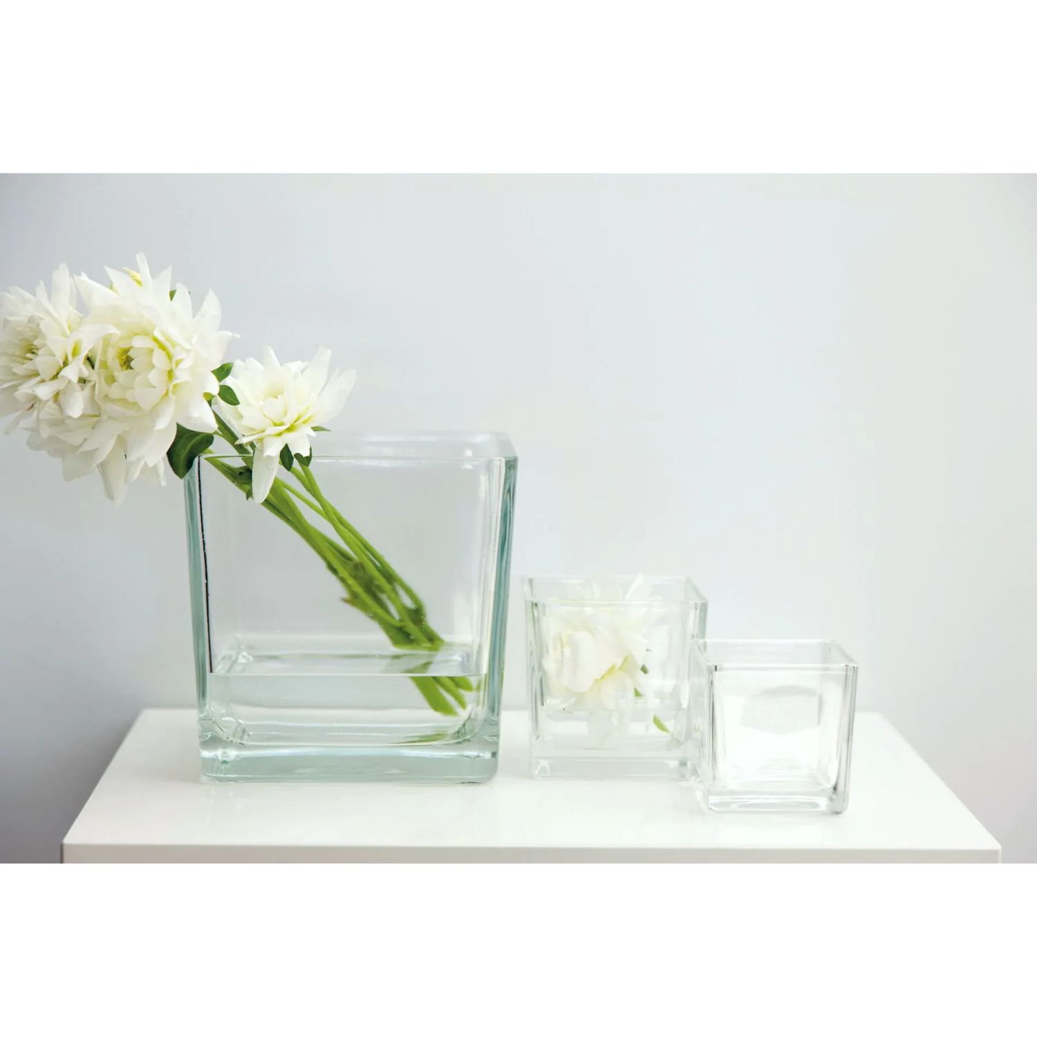 Drei quadratische Glasvasen auf weißer Fläche, links mit weißen Blumen, zwei leer
