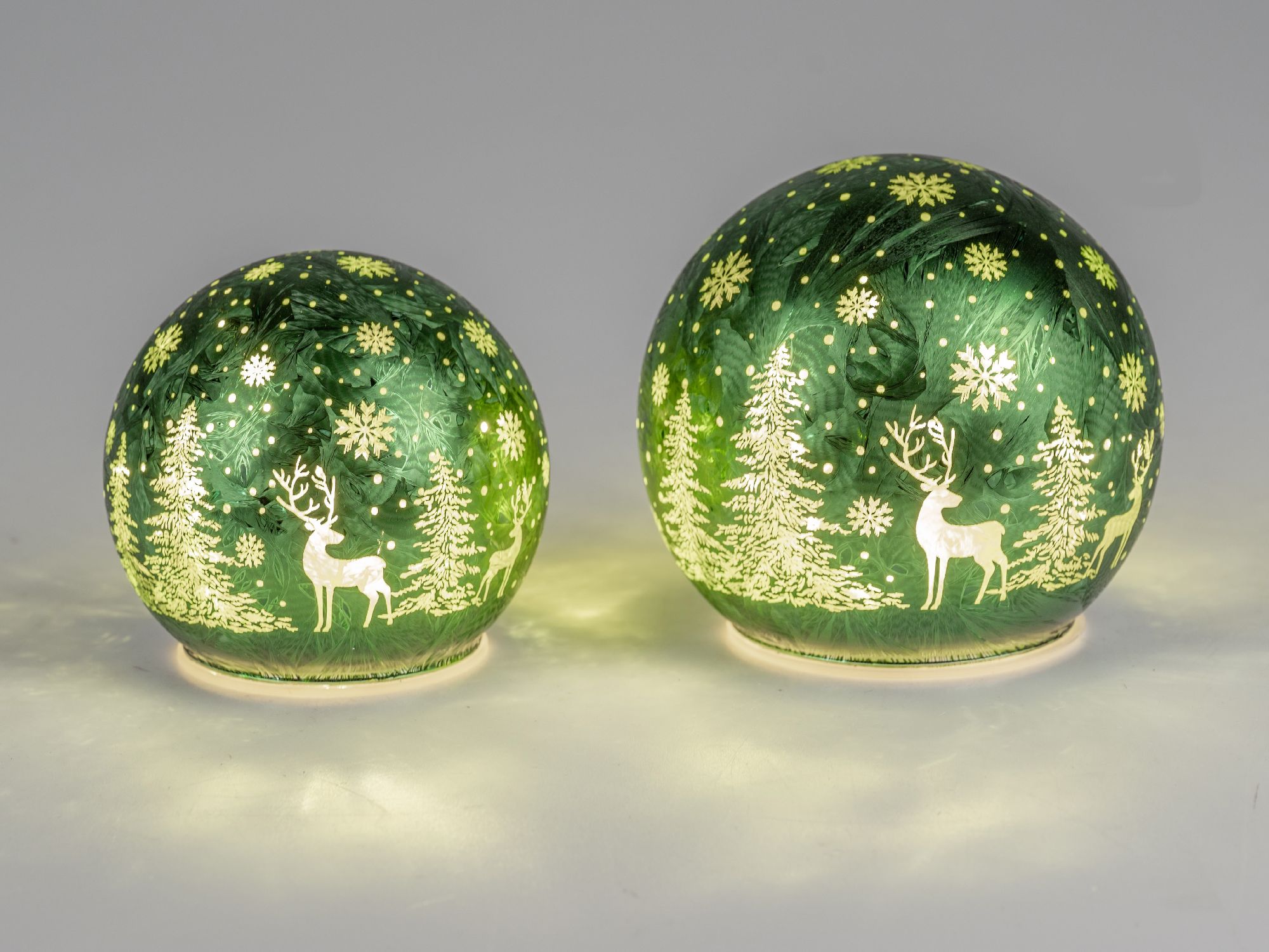 Zwei grüne Glaskugeln mit winterlichen Motiven, darunter Rehe und Tannenbäume, leuchten sanft im Hin