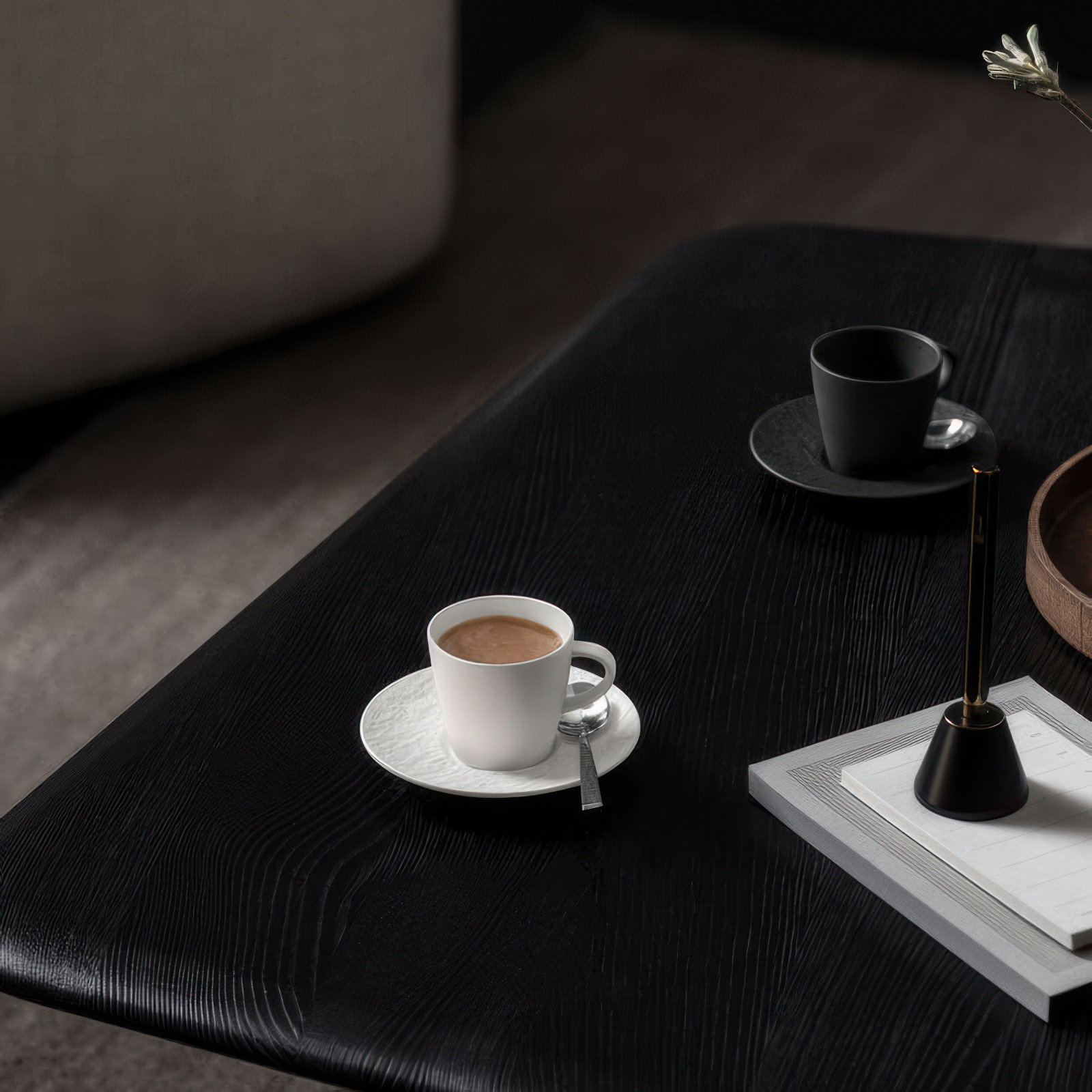 Zwei Kaffeetassen auf einem schwarzen Tisch: eine weiße Tasse mit Untertasse und eine schwarze Tasse. Nebenan ein Notizblock und ein Stift.