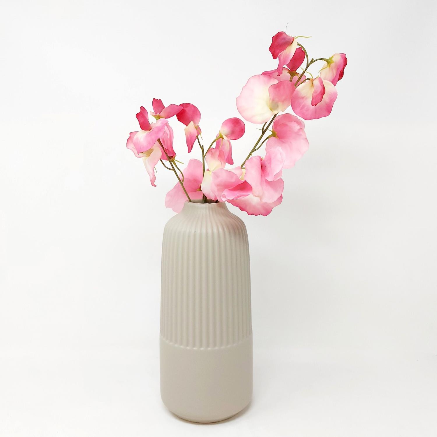Eine beige, gerippte Vase mit pinken Blüten steht vor einem weißen Hintergrund. Die Blüten sind in verschiedenen Rosa-Tönen.