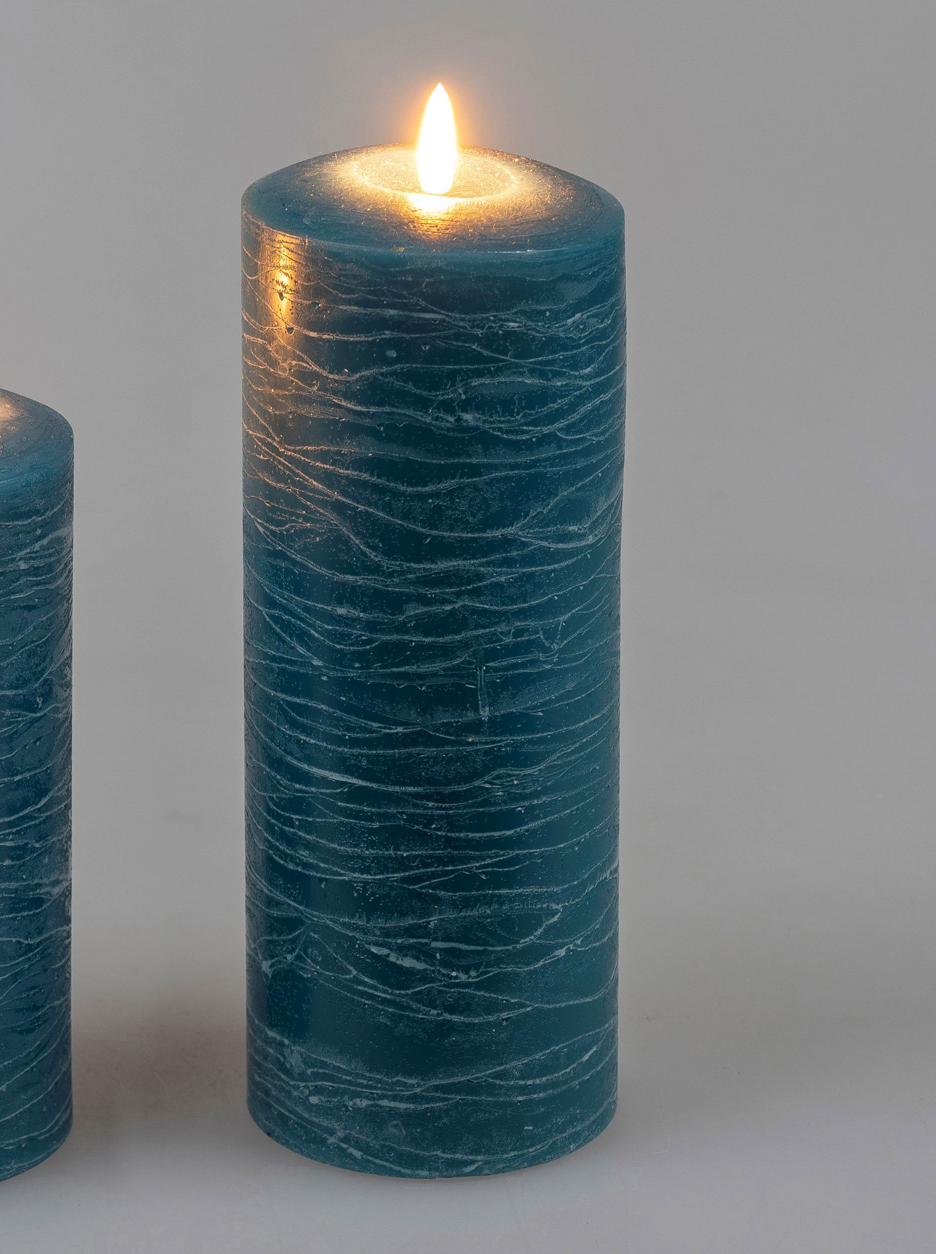 Zwei blaue Stumpenkerzen, eine davon brennt. Die Kerzen haben eine strukturierte Oberfläche und stehen auf einem hellen Hintergrund.