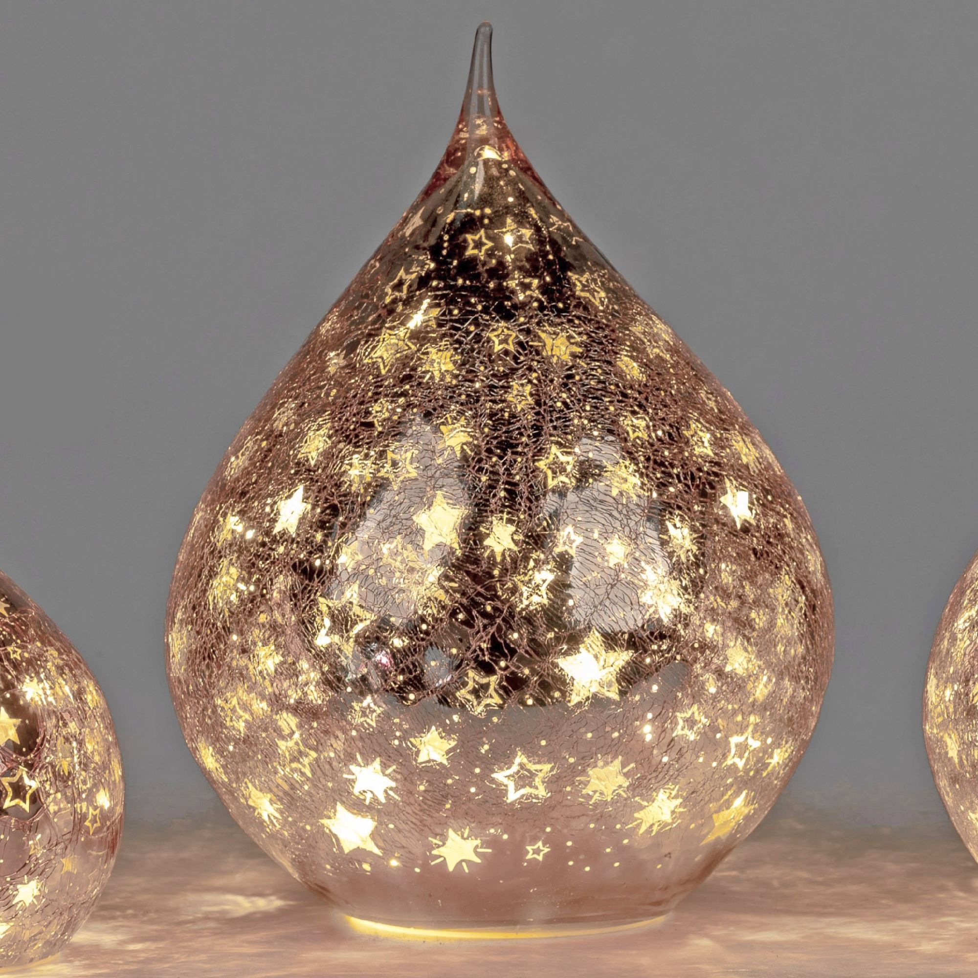 Drei gläserne, tropfenförmige Objekte mit goldenen Sternen und warmem Licht auf grauem Hintergrund.