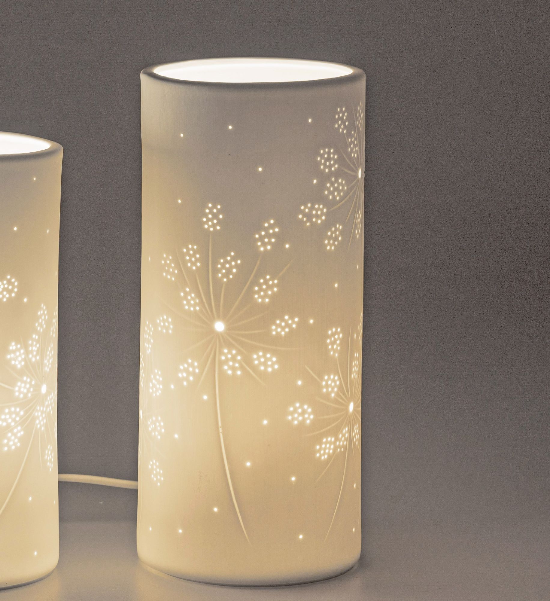 Zwei zylindrische Lampen mit floralem Muster, die sanft leuchten, stehen vor einem grauen Hintergrun
