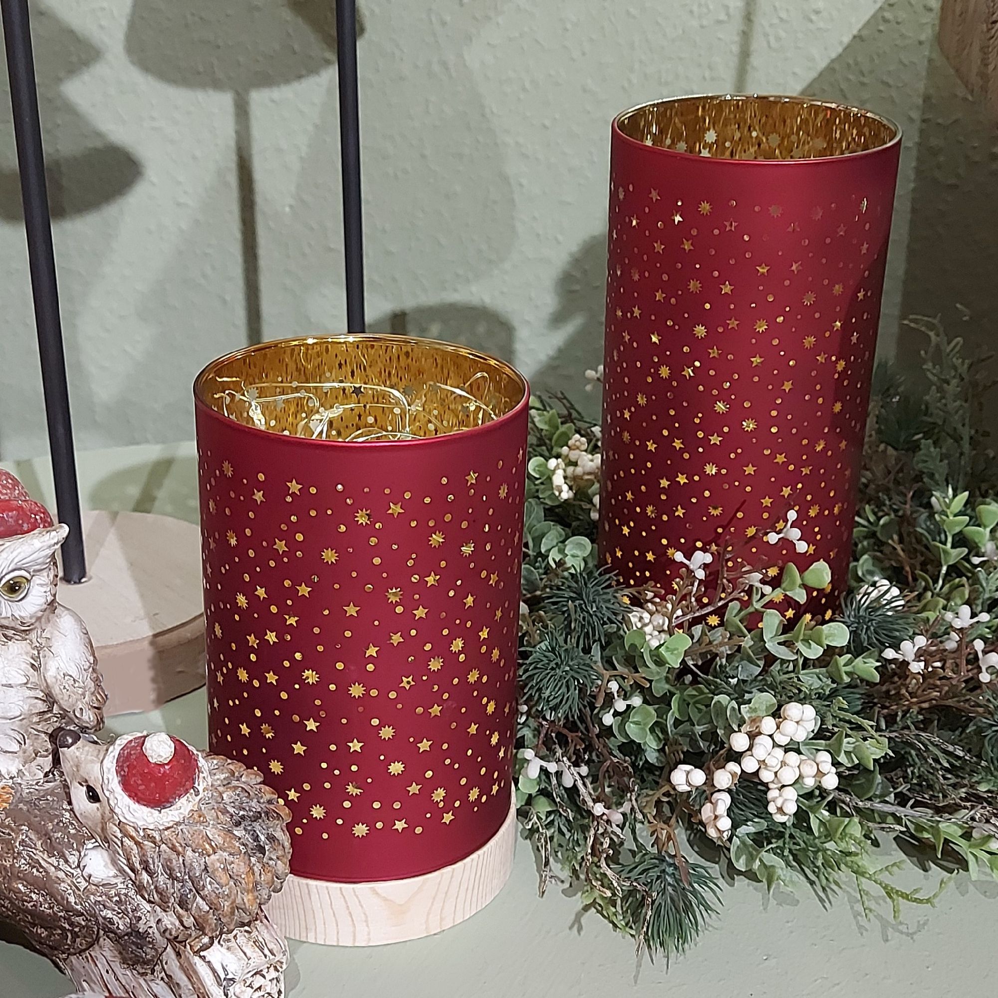 Zwei rote Kerzenhalter mit goldenen Sternen, umgeben von einer grünen Dekoration aus Zweigen und kle