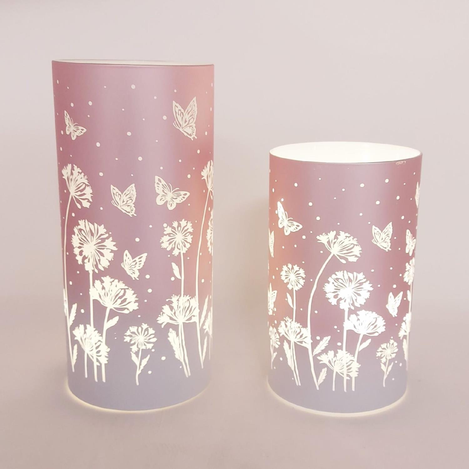 Zwei zylindrische Lampen in sanften Farben mit floralen und schmetterlingsartigen Motiven auf der Oberfläche.