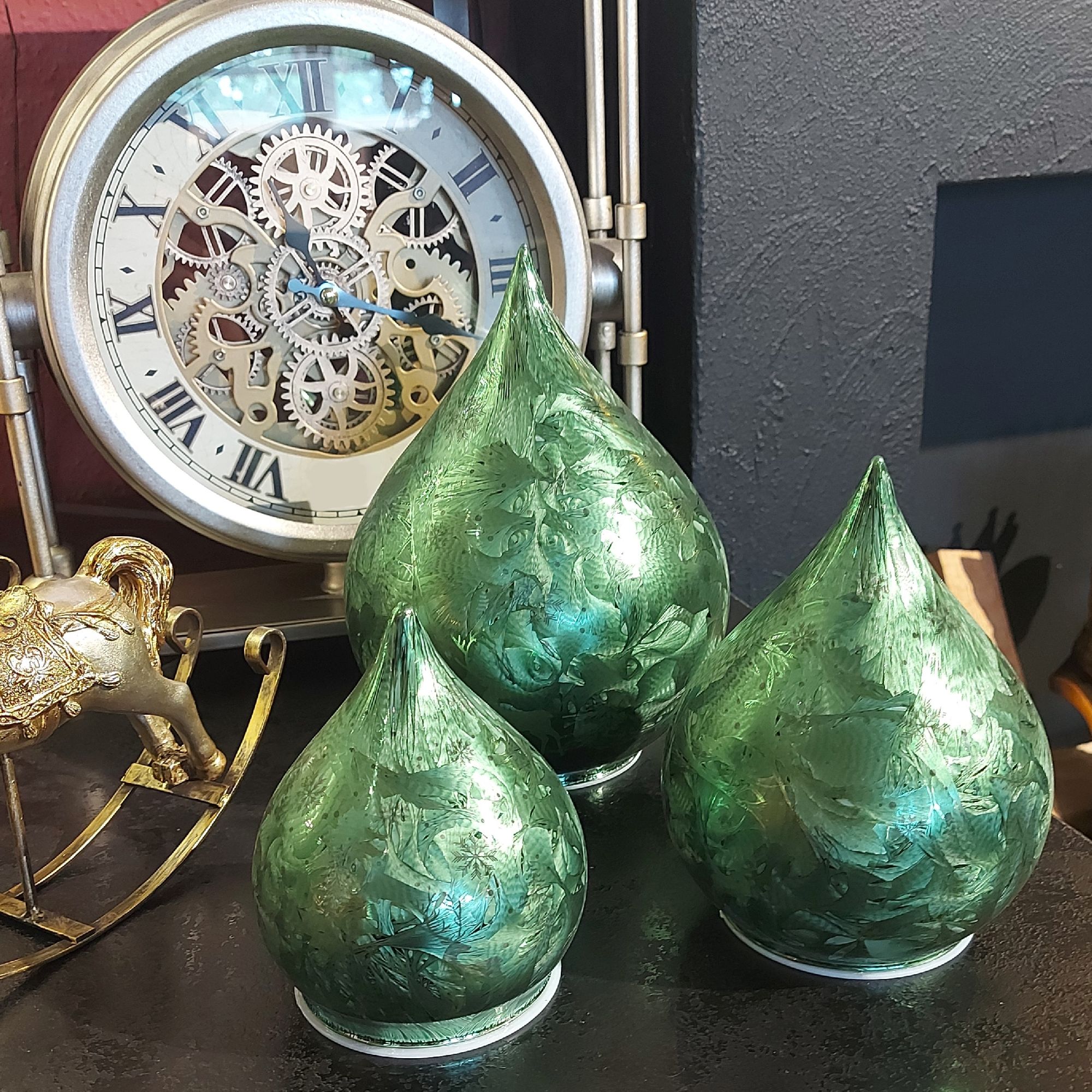 Drei grüne, tropfenförmige Objekte mit floralem Muster stehen vor einer Uhr mit sichtbaren Zahnräder