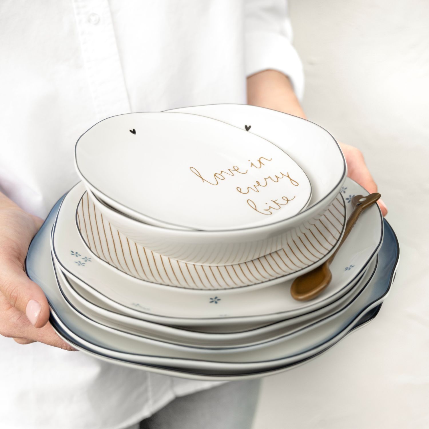 Hände halten Stapel weißer Teller und Schalen mit Goldlöffel und Schriftzug „Love in every bite“