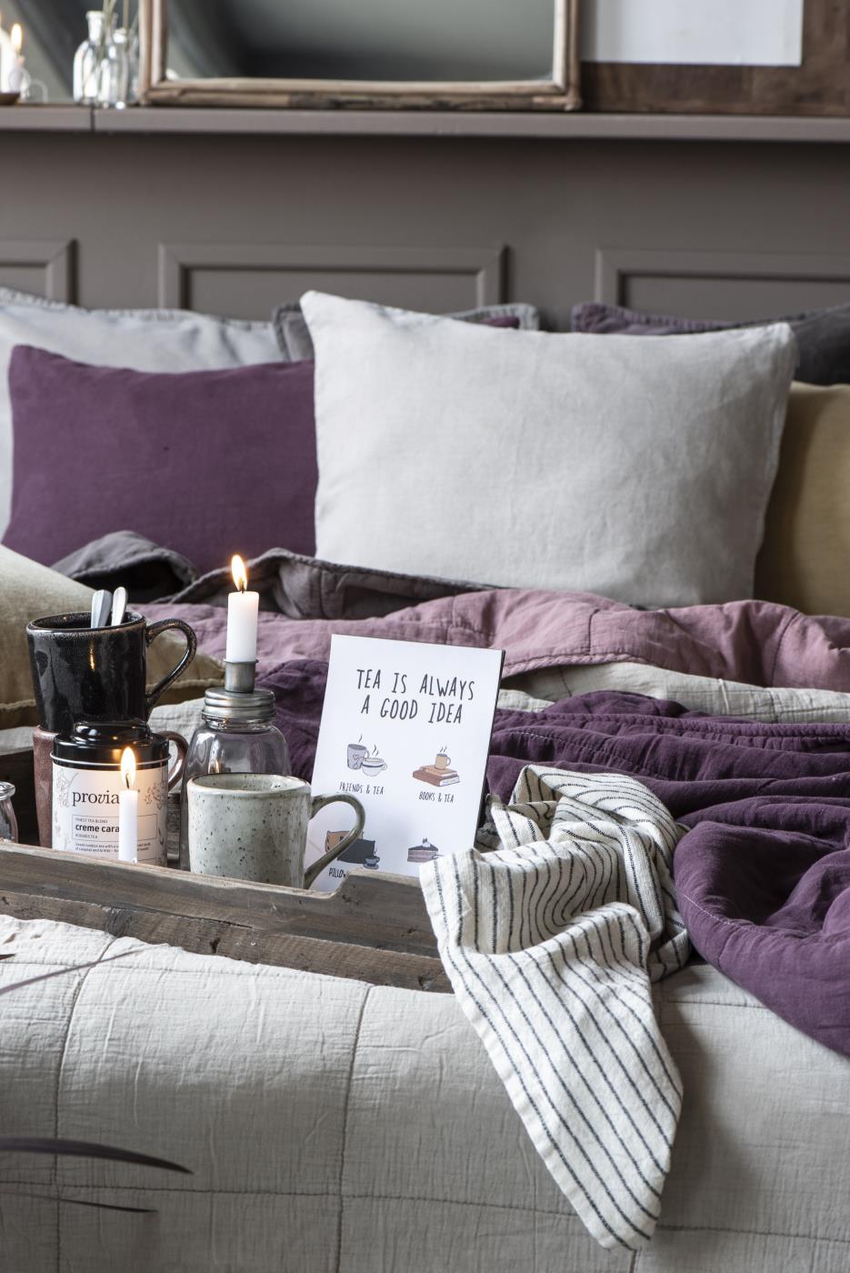 Ein Bett mit bunten Kissen, einer Decke, einer Holzplatte mit Teetassen, einer Kerze und einem Schild mit der Aufschrift „Tea is always a good idea“.