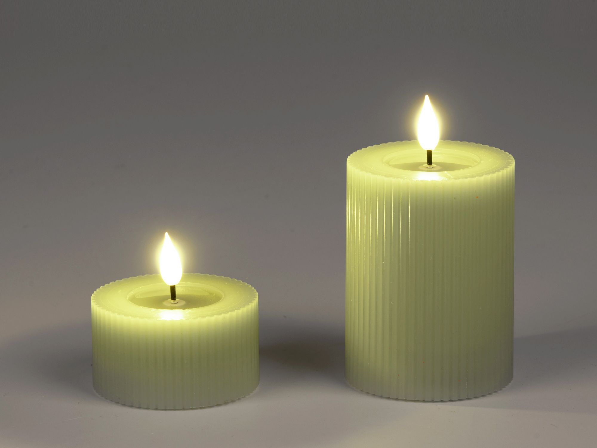 Zwei grüne Kerzen stehen auf einem neutralen Hintergrund. Die größere hat eine gerade Form, die klei