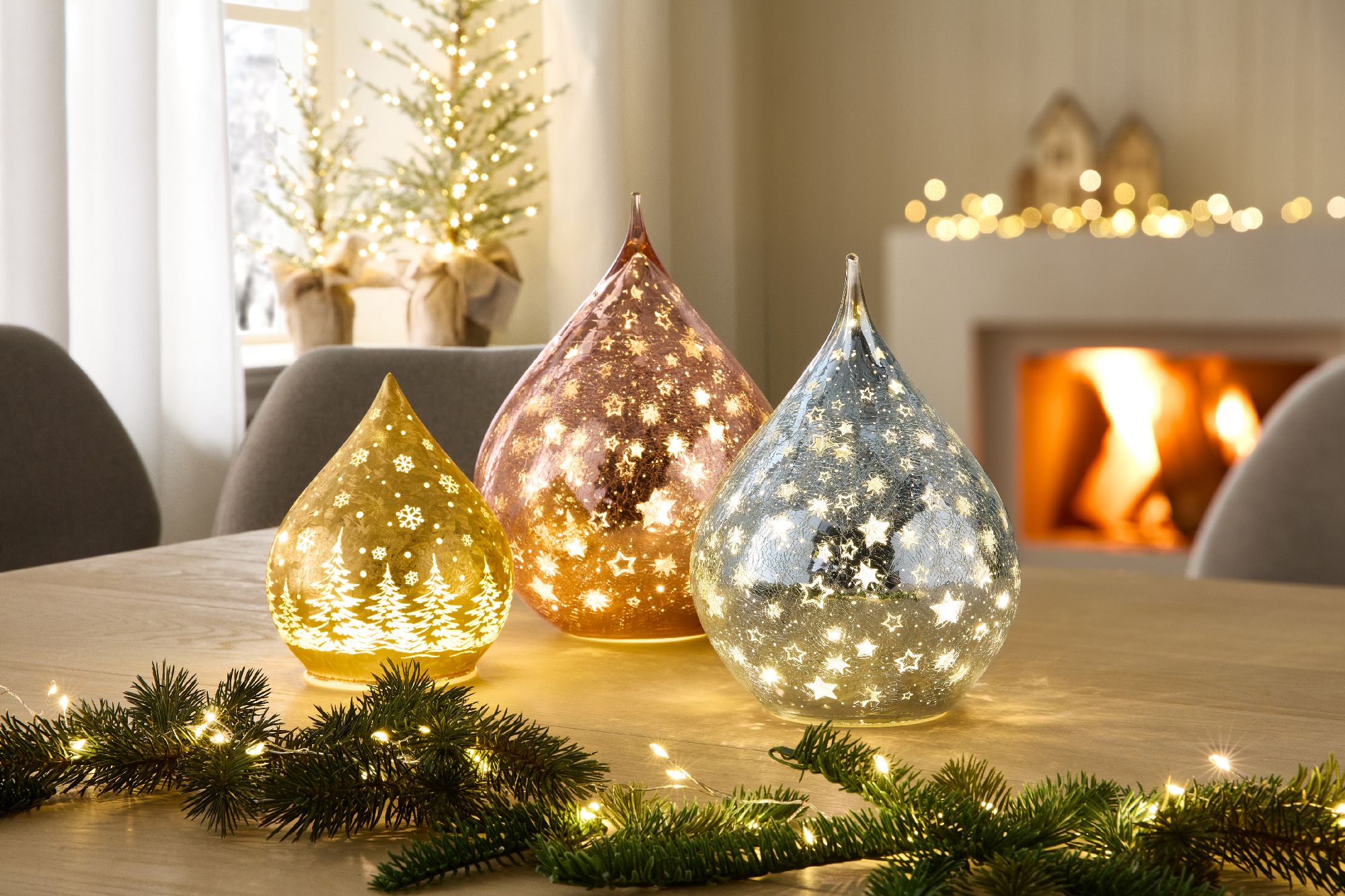 Drei leuchtende, tropfenförmige Dekolichter auf einem Tisch, umgeben von Tannenzweigen, im Hintergru