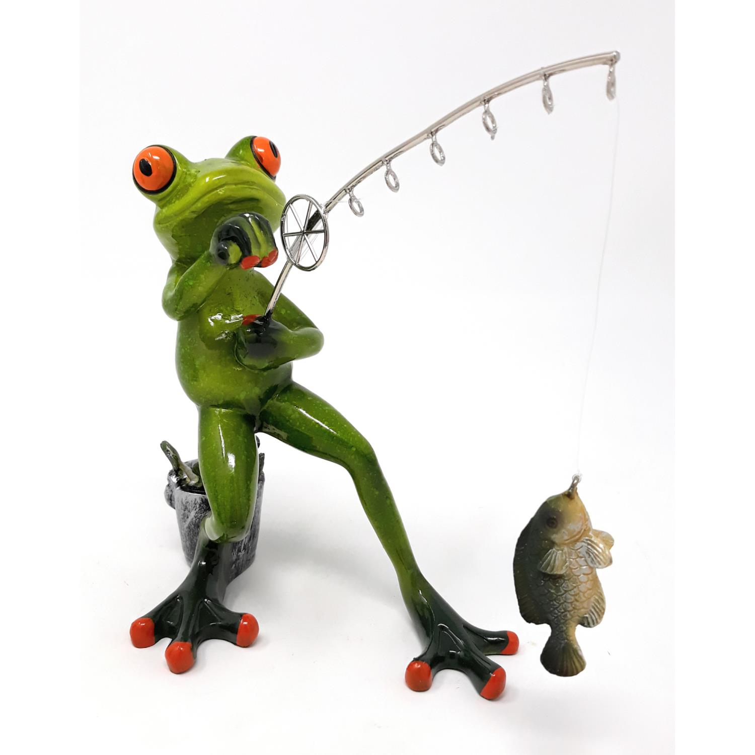 Eine grüne Froschfigur sitzt auf einem Eimer und angelt mit einer Rute, an der ein kleiner Fisch hängt.