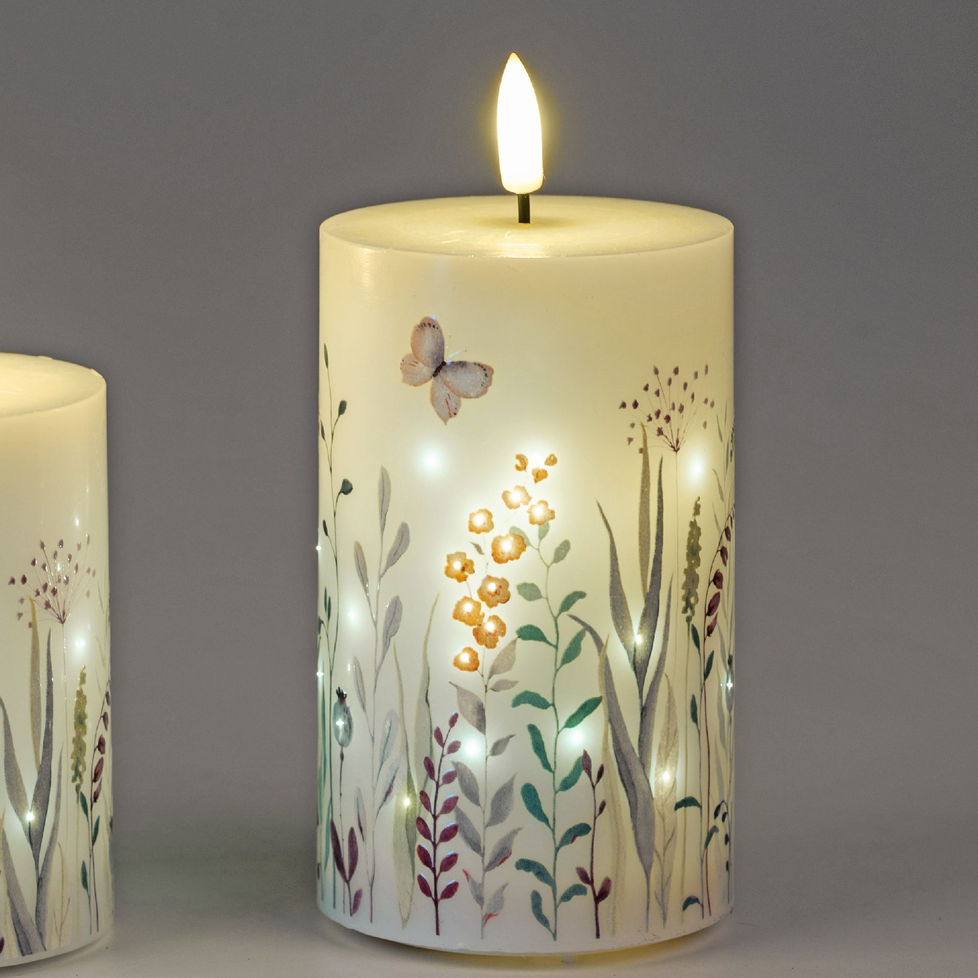 Zwei Kerzen mit floralen Motiven, eine brennend, die andere unbeleuchtet. Eine Schmetterlingsillustr