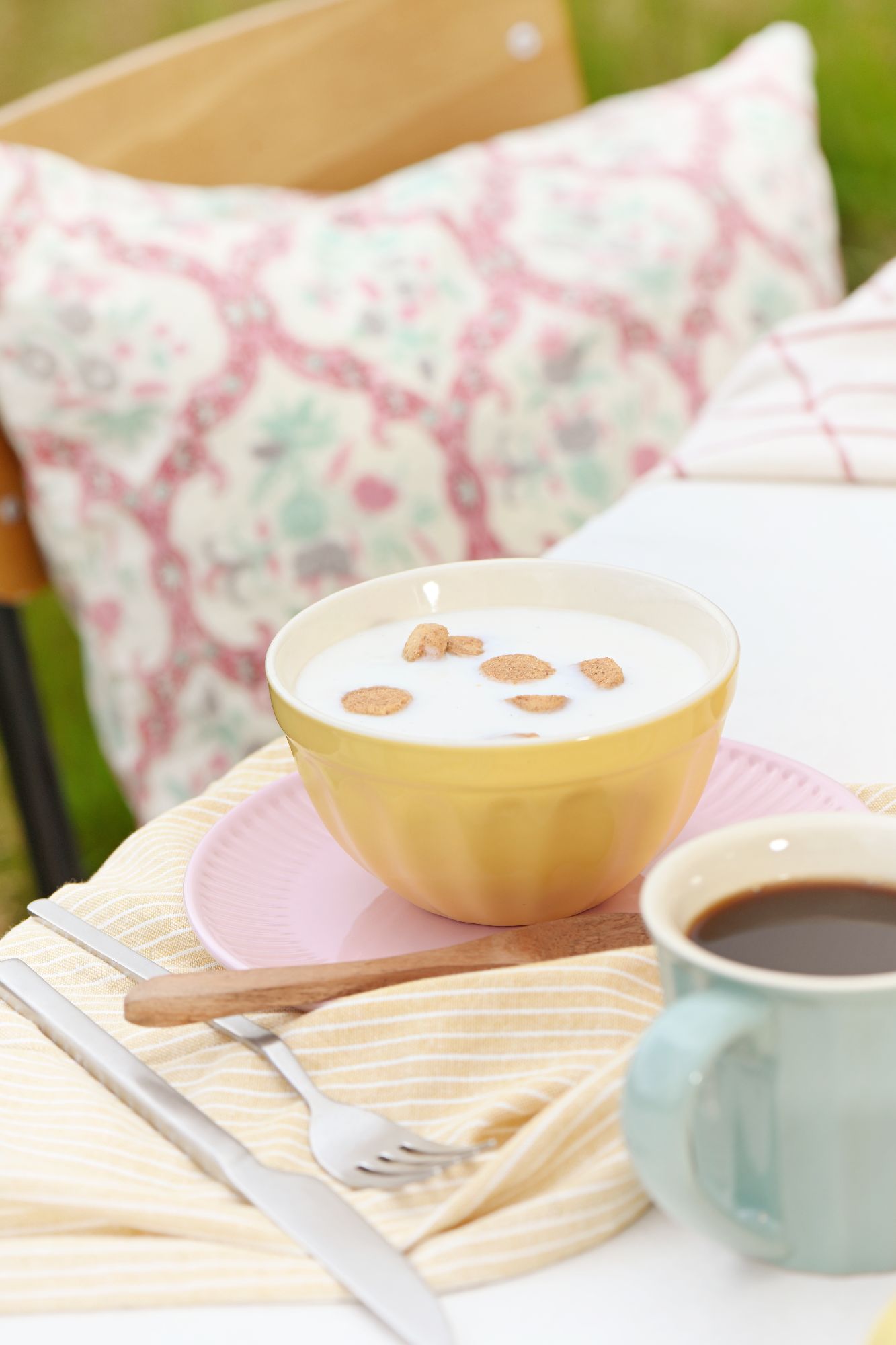 Eine gelbe Schüssel mit Milch und Cerealien steht auf einem rosa Teller. Daneben eine Tasse Kaffee und Besteck auf einem gestreiften Tuch.