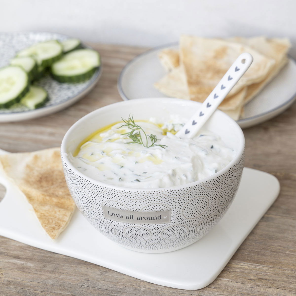 Eine Schüssel mit Joghurt, garniert mit Dill und Olivenöl, steht auf einem Tisch. Daneben liegen Gurkenscheiben und Fladenbrot.