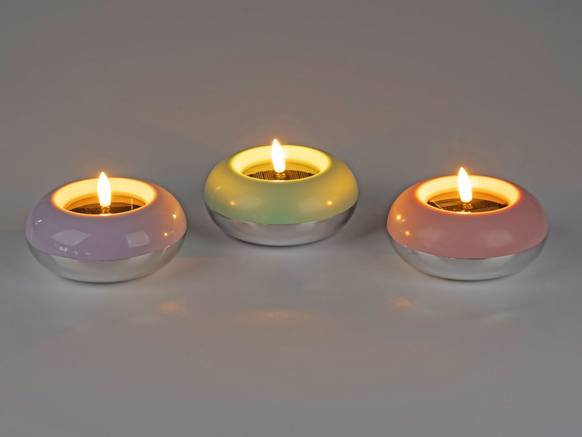 Drei runde Teelichthalter in lila, grün und rosa mit silbernem Boden, jeweils mit einer brennenden Kerze.