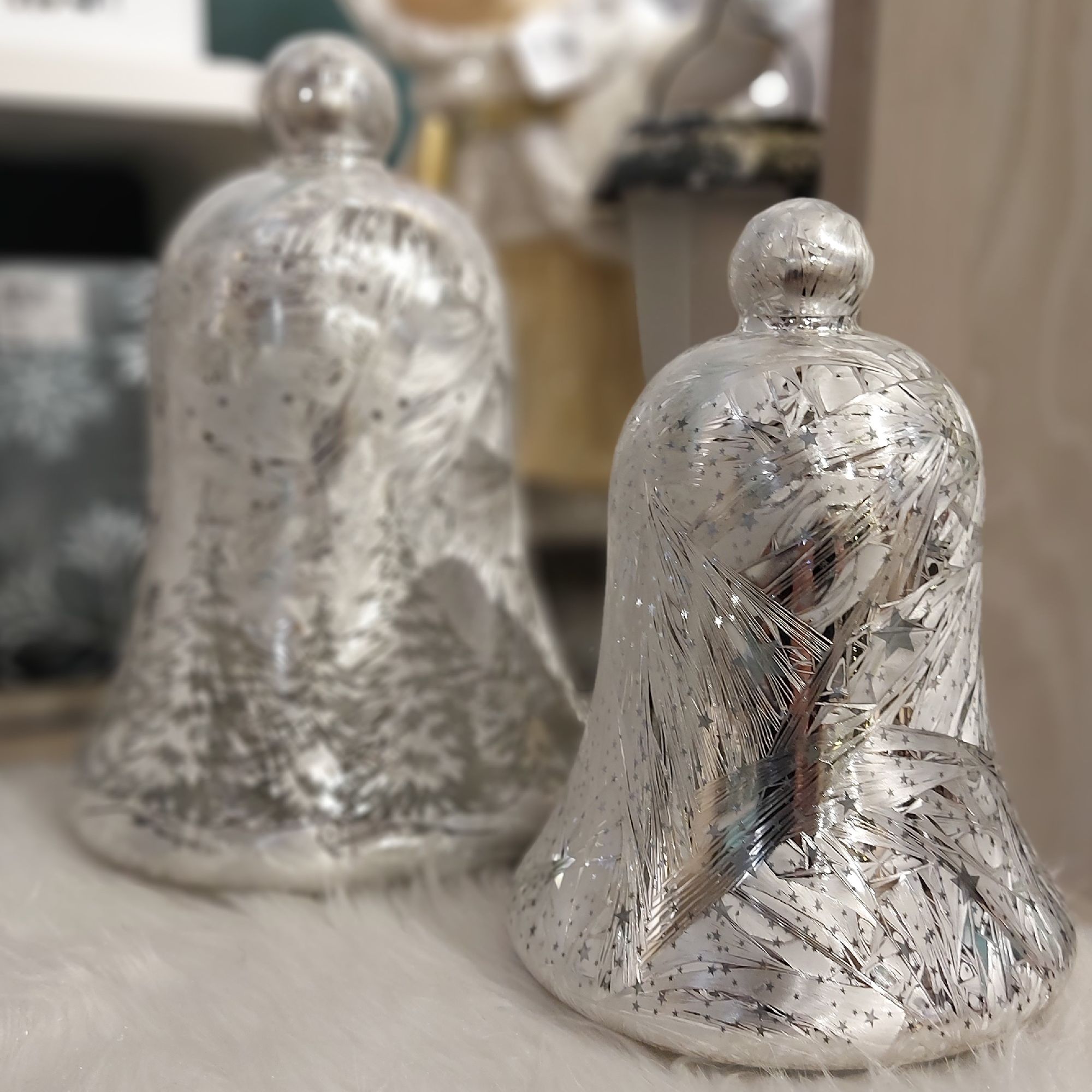 Zwei silberne Glöckchen mit glitzernden Mustern stehen auf einem weißen Untergrund. Im Hintergrund s
