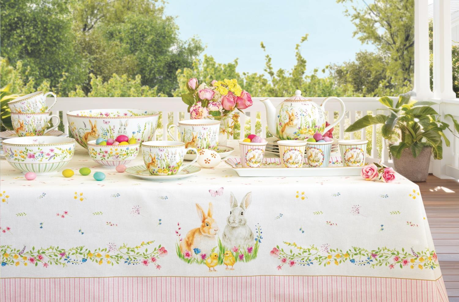 Tischgedeck mit bunten Tellern, Tassen, einer Teekanne und Dekoration aus Blumen und Ostereiern auf einem Tisch mit Tischdecke.