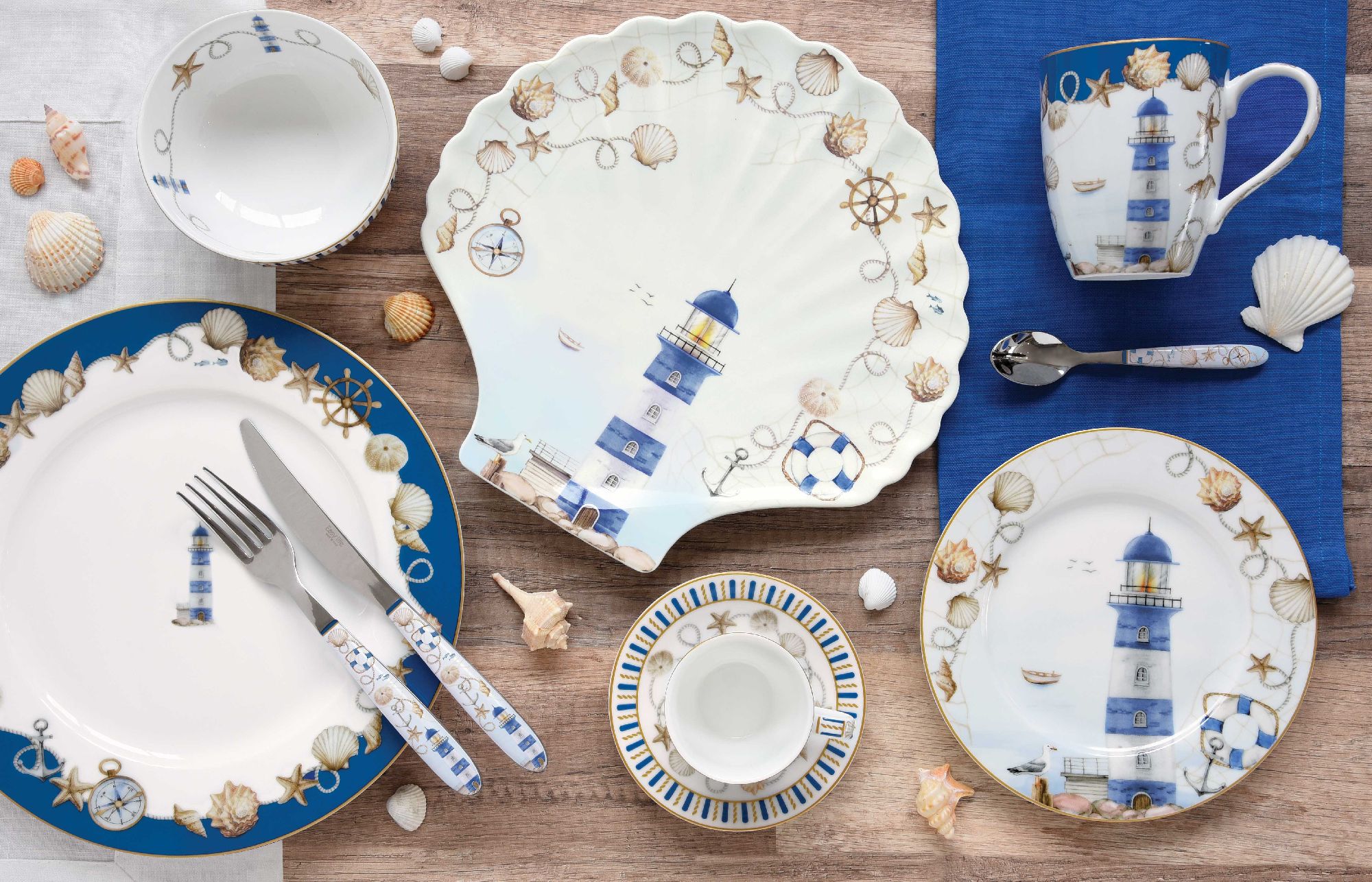 Tischgedeck mit maritimem Design: Teller, Schalen und Tasse mit Leuchtturm-Motiven, umgeben von Muscheln und Dekoration.