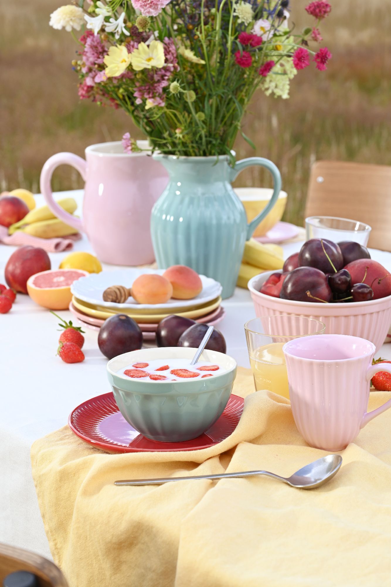 Ein gedeckter Tisch im Freien mit bunten Tellern, Schalen, Gläsern, Obst und einem Blumenstrauß in einer Vase.